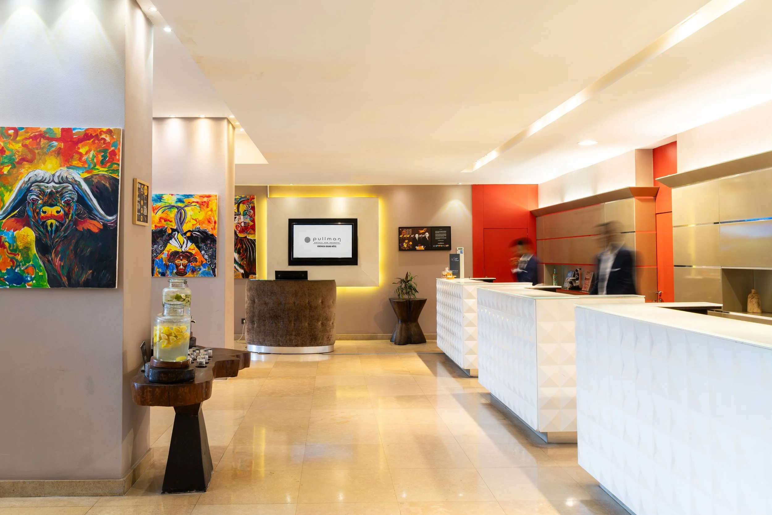 Réalisation moderne de la réception d'un hôtel ou bureau avec des peintures colorées accrochées et deux réceptionnistes en arrière-plan, floues, derrière le comptoir blanc.