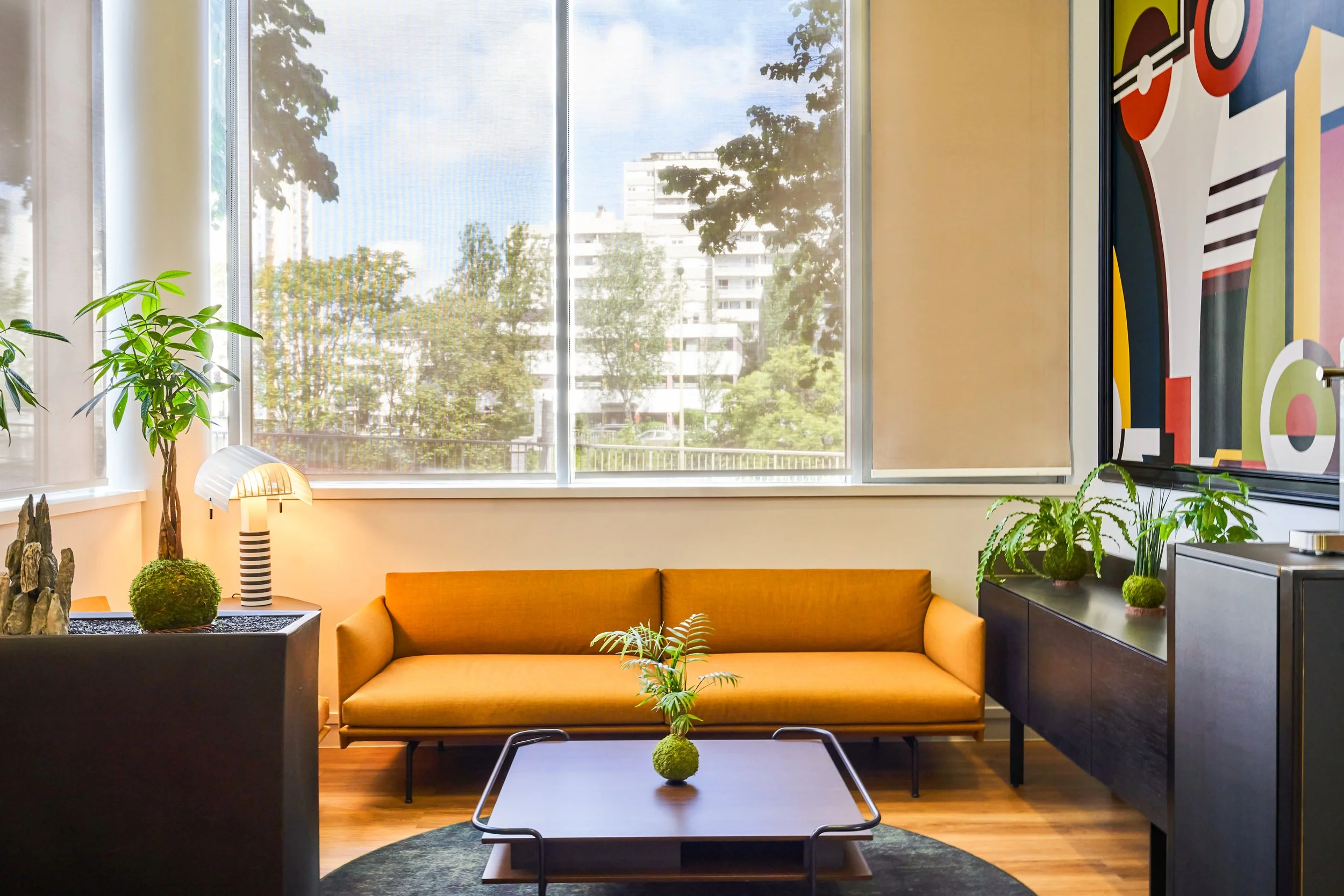 Salon moderne avec un canapé orange, une table basse avec une plante verte, plusieurs plantes d'intérieur, un mur avec une peinture abstraite, grande fenêtre laissant entrer la lumière naturelle.