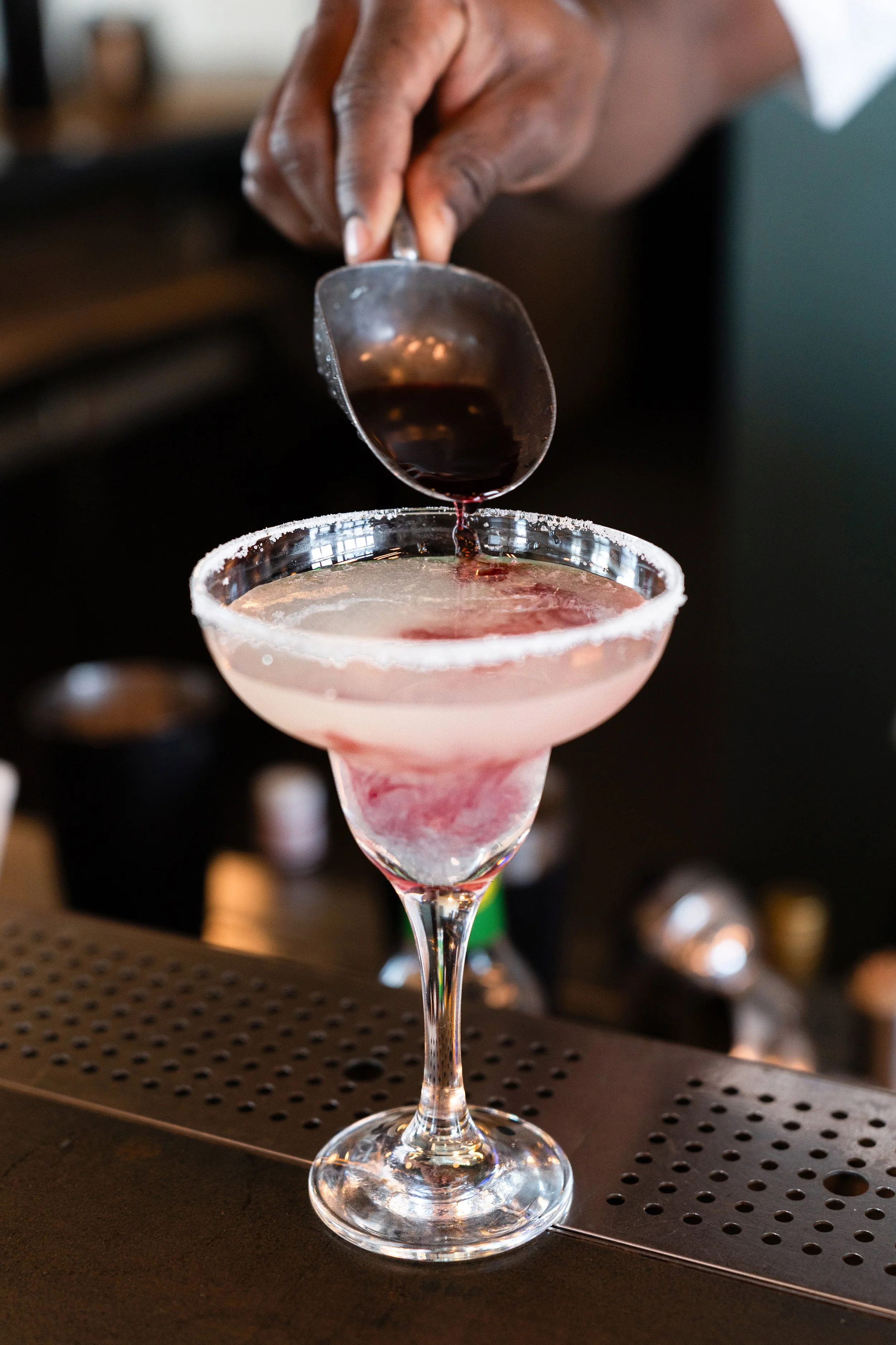 Un barman verse une boisson dans un verre à cocktail décoré du bord en sel, sur un comptoir de bar.