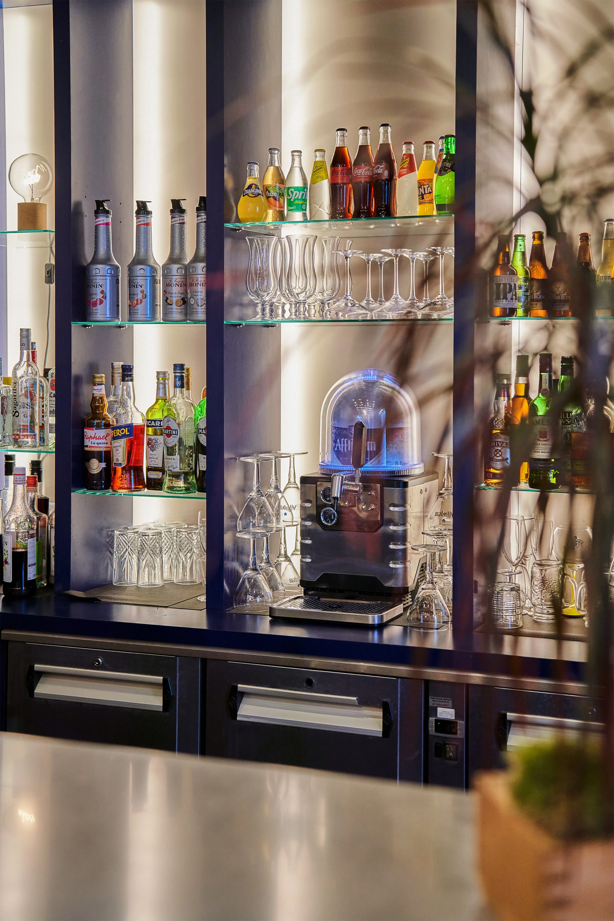 étagère de bar avec bouteilles de boissons gazeuses et alcoolisées, verres à pied, une machine à café, un éclairage modulé, et un comptoir en métal.