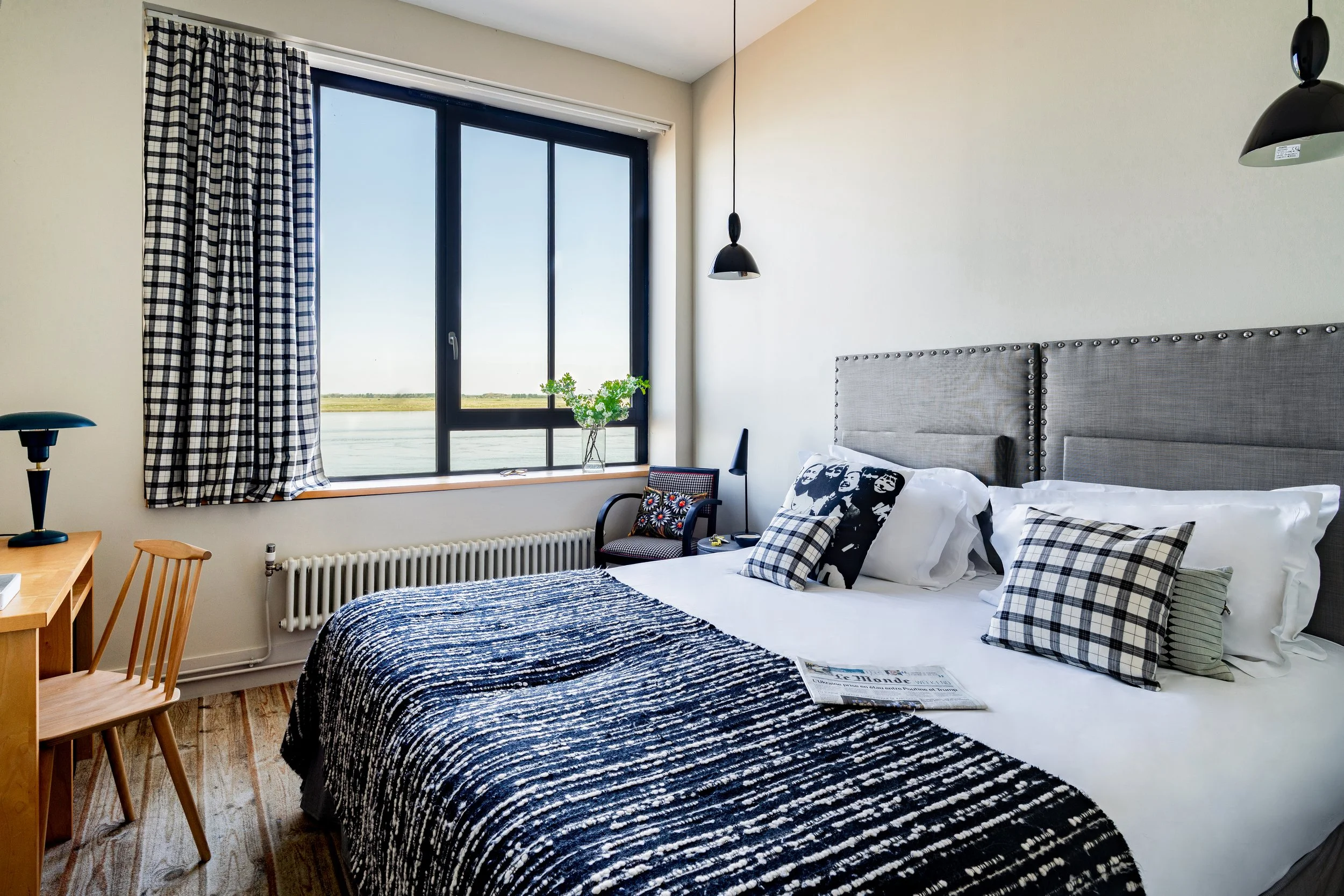 Chambre de style moderne avec un grand lit, des coussins à motifs à carreaux, une fenêtre donnant sur un paysage d'eau, un fauteuil, une table de chevet avec une lampe noire, et des rideaux à carreaux noirs et blancs.