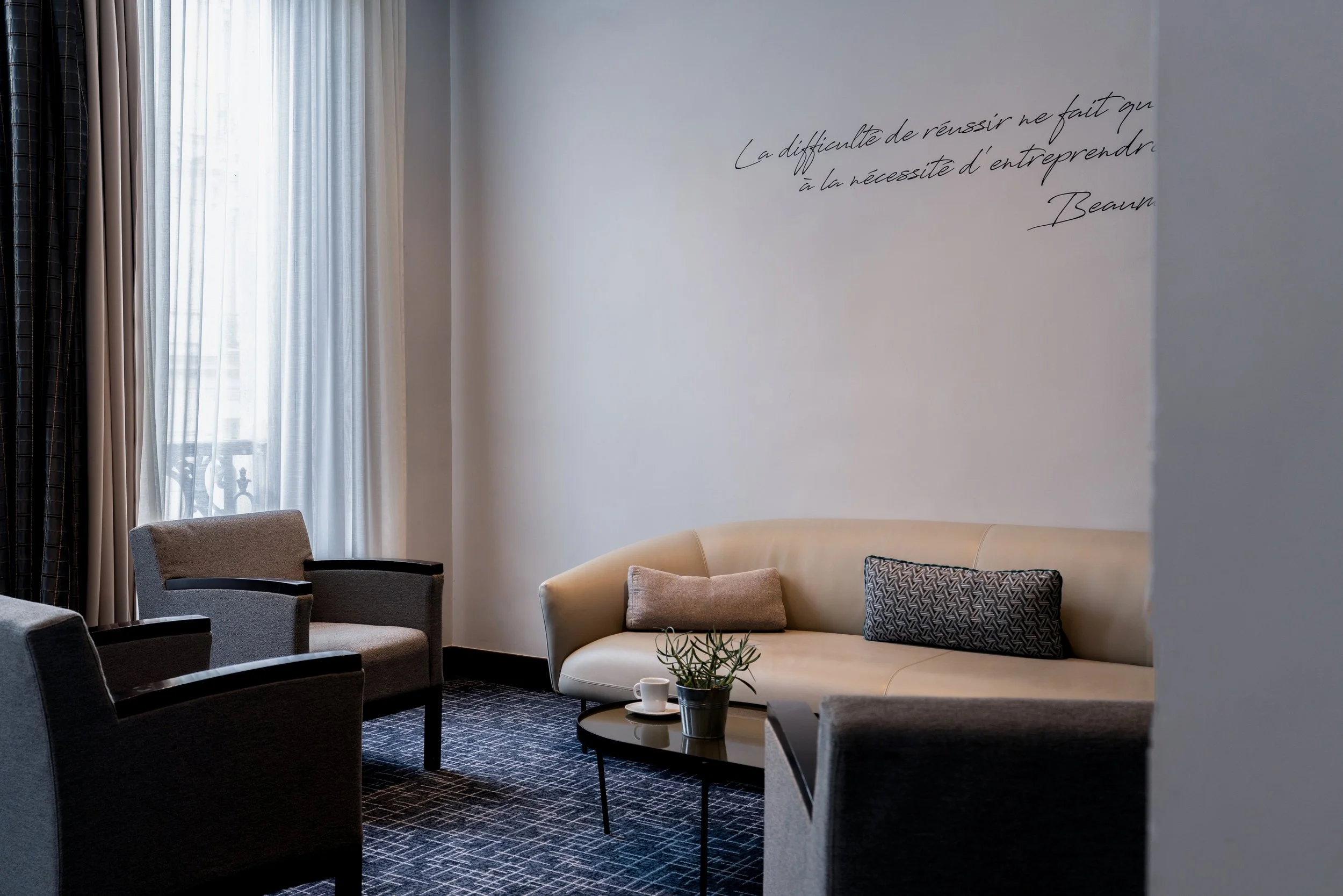 Salon avec trois fauteuils, un canapé beige, une table basse avec une plante, tasse, et coussins, devant une fenêtre avec des rideaux, mur avec citation en français.