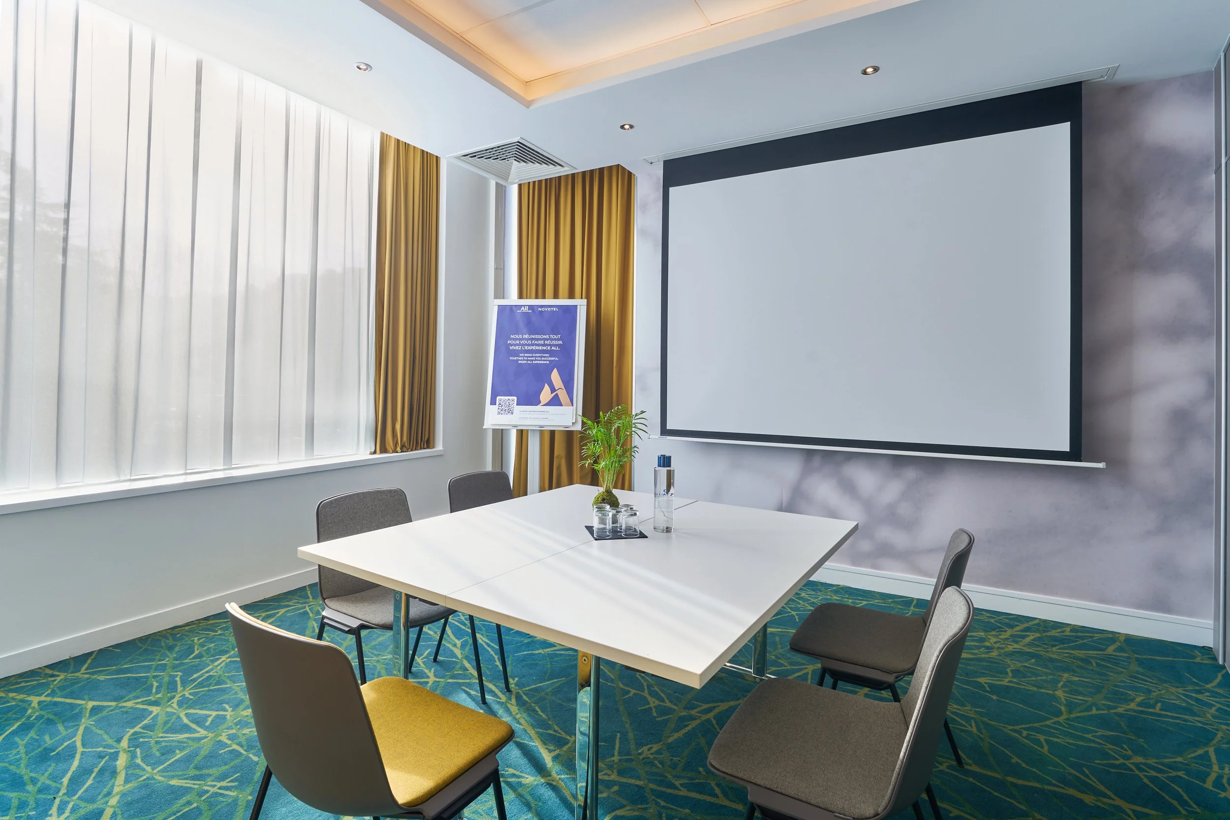 Salle de réunion en style boardroom avec une grande table blanche, six chaises, un écran de projection, un grand écran, et des fenêtres avec rideaux blancs et dorés.