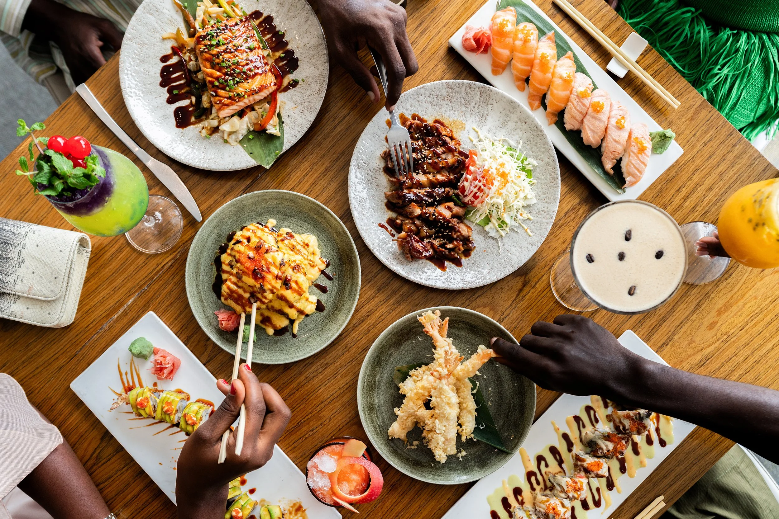 Groupe de personnes partageant un repas japonais avec sushis, sashimis, tempura, et boissons colorées sur une table en bois.