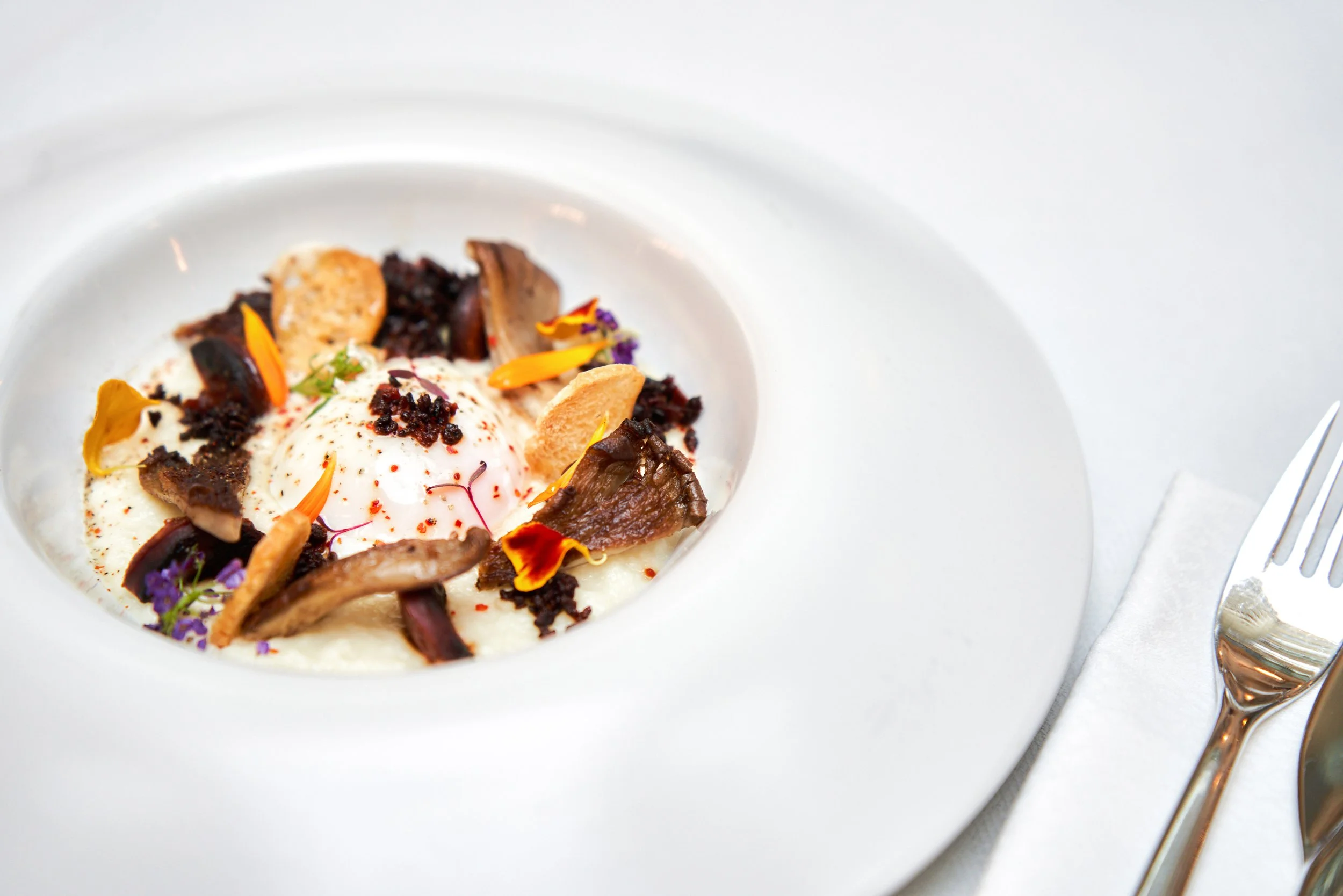 Plat gastronomique avec œuf poché, champignons, fleurs comestibles, chips et sauce, présenté dans une assiette blanche haut de gamme.
