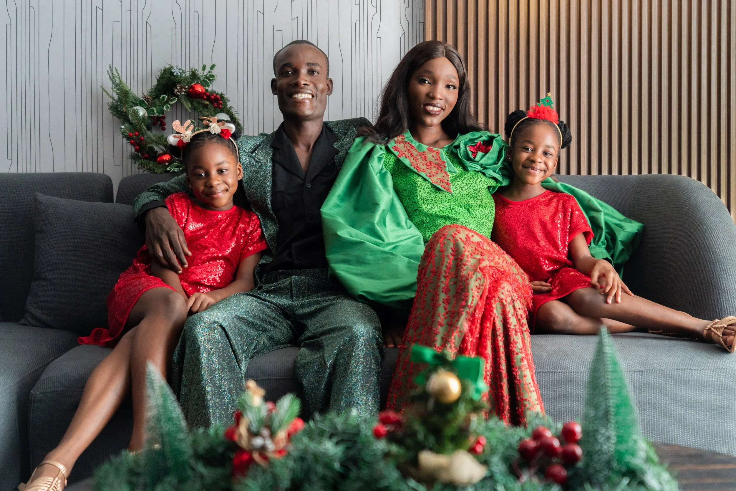 Une famille en tenue de fêtes, assise sur un canapé, devant une décoration de Noël, comprenant un gros guirlande verte avec des ornements rouges.