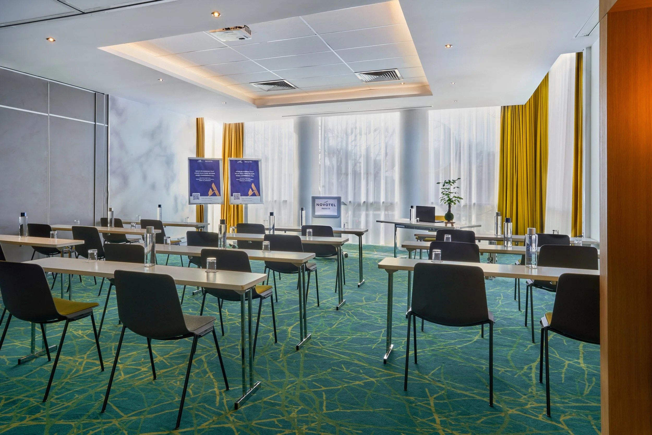 Salle de conférence en style salle de classe avec des tables et des chaises et un décor lumineux avec des rideaux jaunes et une moquette verte.