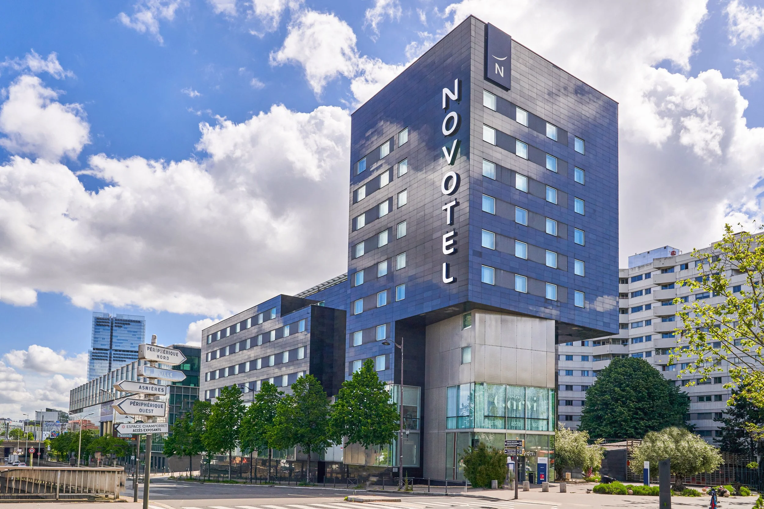 Gratte-ciel moderne avec un panneau lumineux indiquant "NOVOTEL" sur la façade, devant un ciel partiellement nuageux, avec des arbres en premier plan et d'autres bâtiments en arrière-plan.