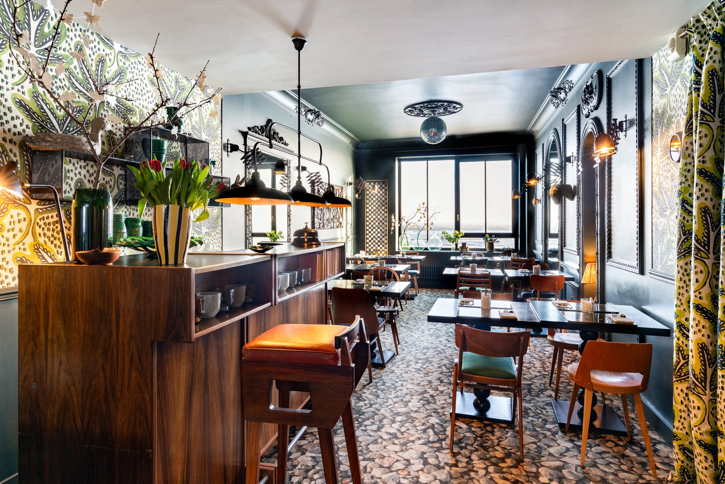 Intérieur d'une salle de petit déjeuner stylée avec mur en papier peint coloré, grandes fenêtres, tables avec assiettes et verres, chaises en bois, décorations florales et éclairage chaleureux.