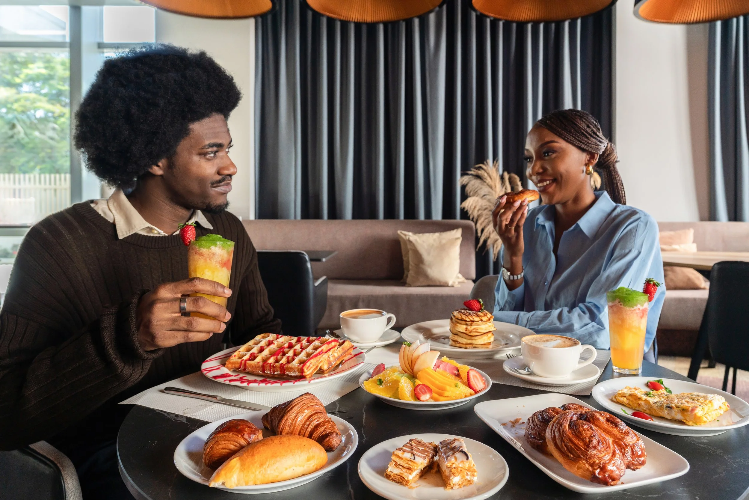 Deux personnes dégustant un petit déjeuner composé de pâtisseries, fruits, cafés, et boissons colorées dans un café accueillant.