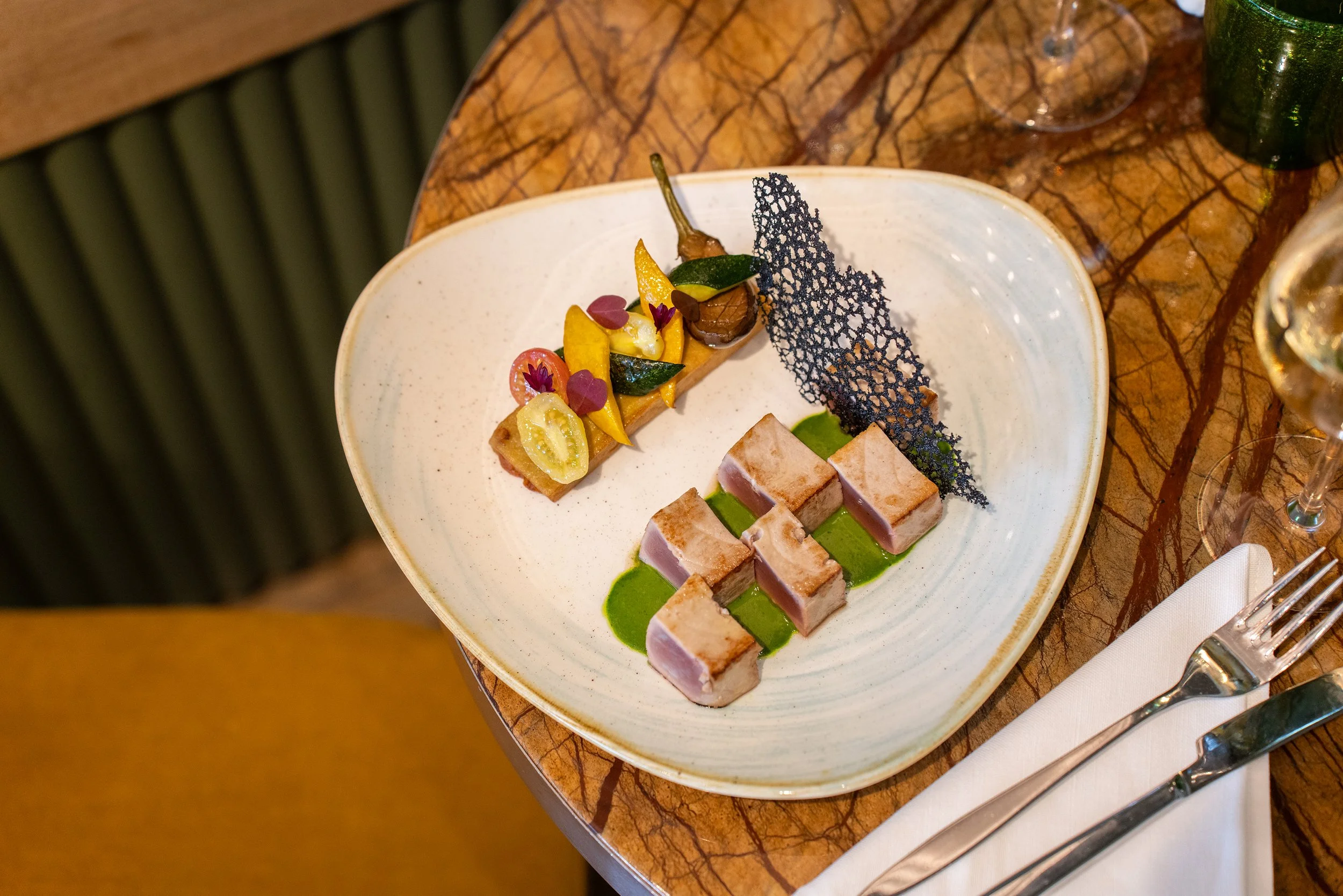 Assiette de gastronomie avec des morceaux de viande ou poisson cuits, accompagnée de légumes colorés et d'une fine tranche de pain ou biscuit, décorée avec une fine feuille de croustillant noir et quelques petits éléments végétaux.