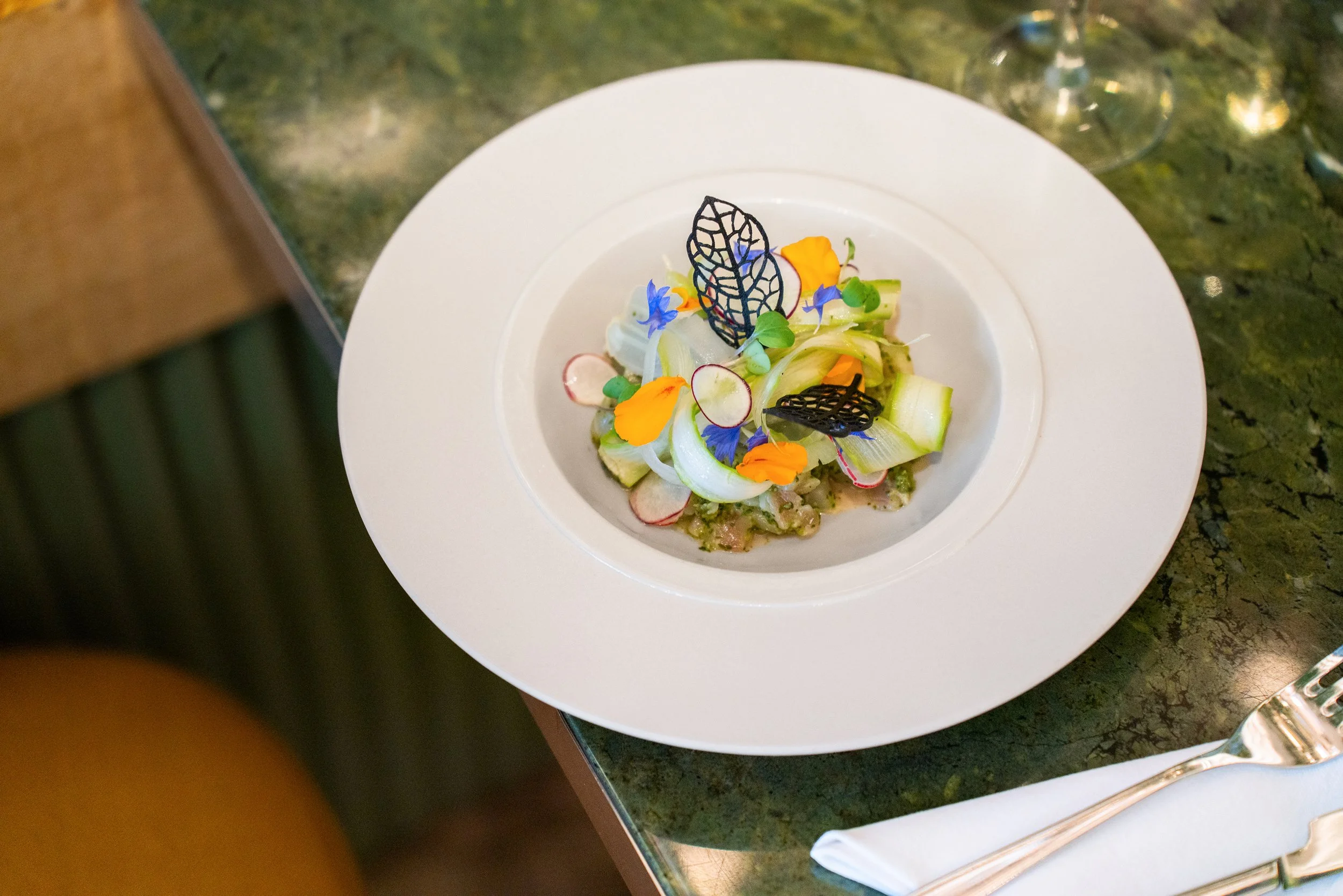 Salade sophistiquée avec des fines rondelles de radis, de la laitue, des fleurs comestibles, et des décorations en feuille noire, présentée sur une assiette blanche sur une table en marbre vert.