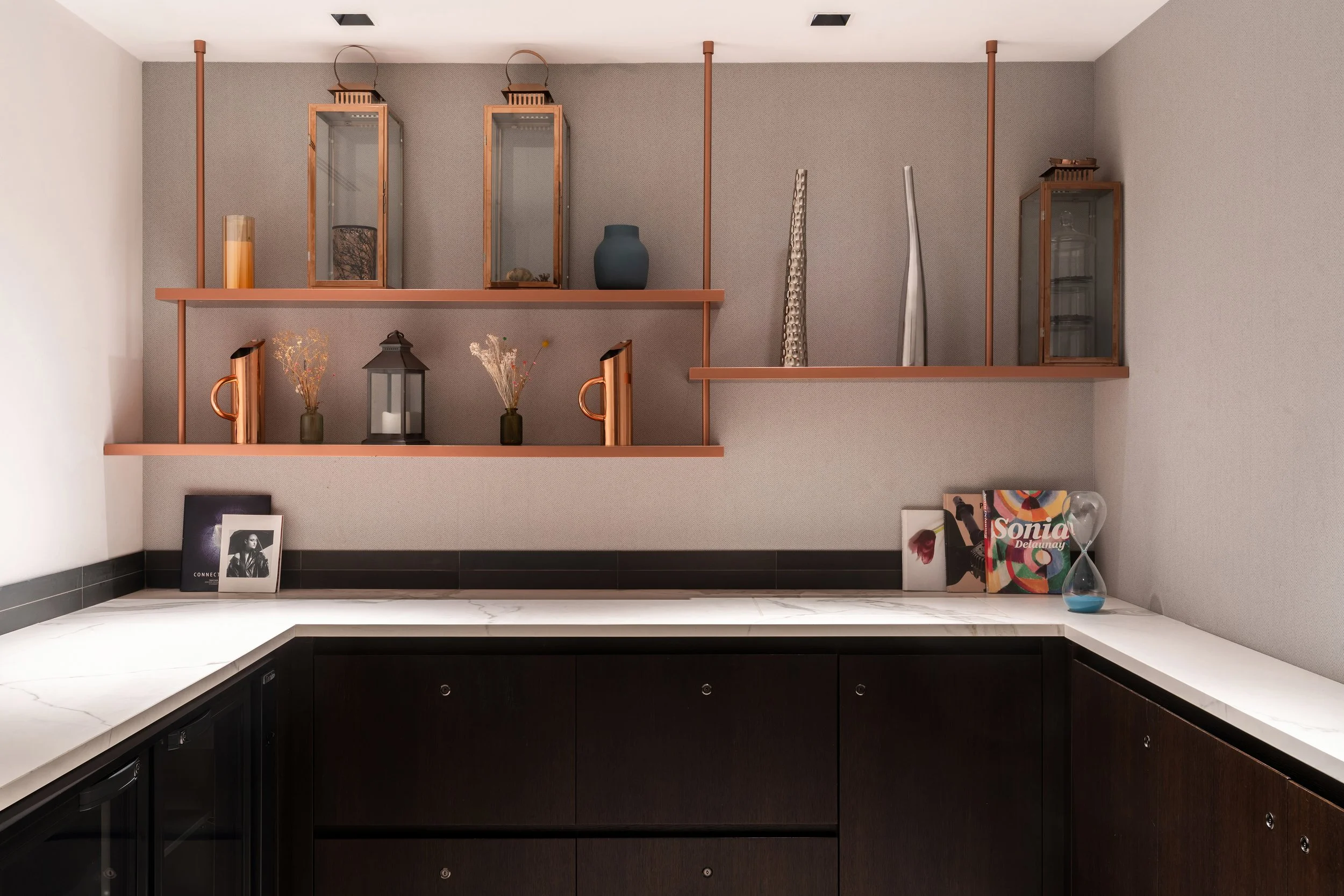 Étage décoré au dessus d'un buffet moderne avec étagères en cuivre, vases, lanternes, livres et un sablier, devant un mur gris clair et un plan de travail en marbre noir et blanc.