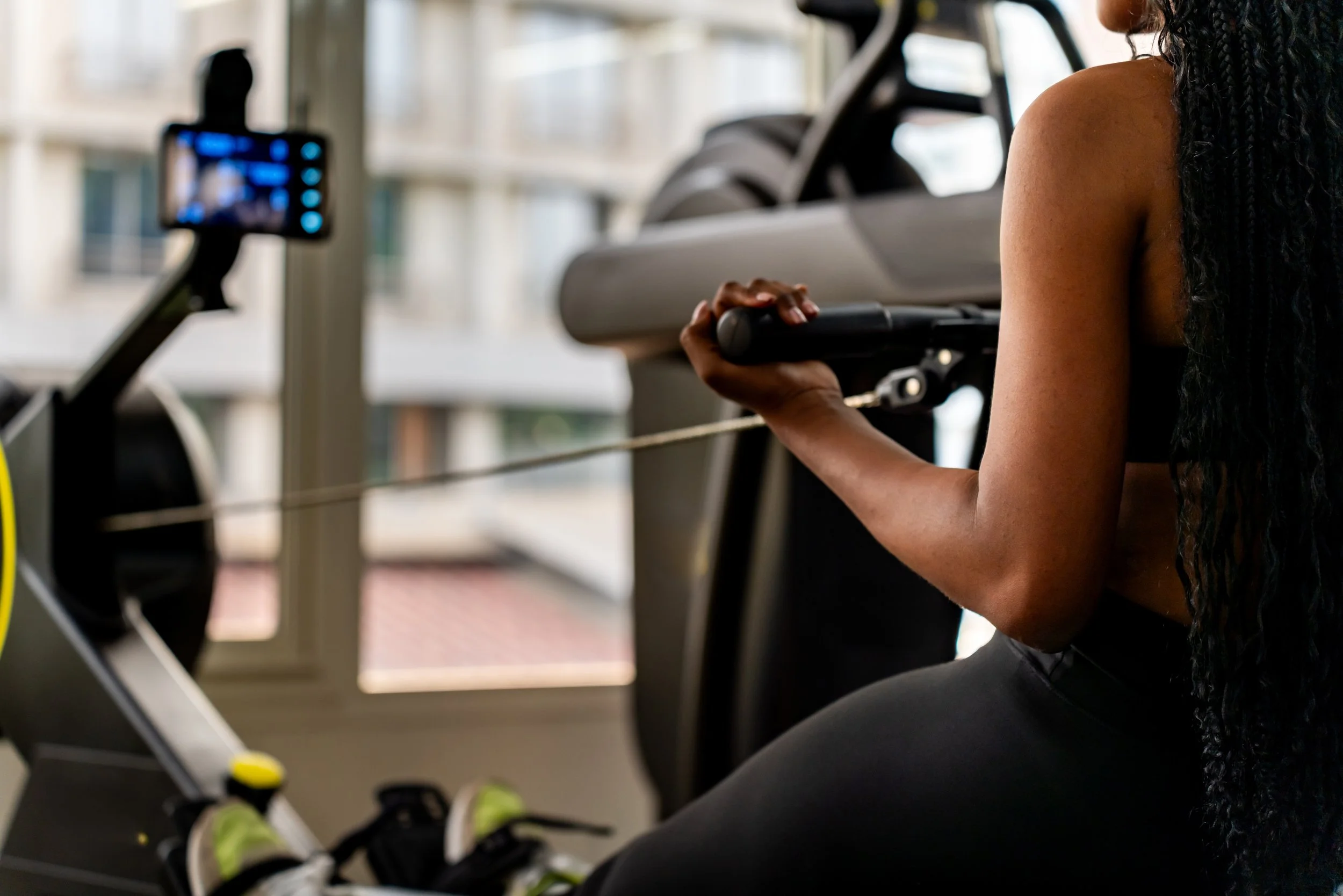 Femme utilisant un rameur d'intérieur, vue de côté, dans une salle avec fenêtres, vêtements de sport noirs et longue chevelure bouclée.