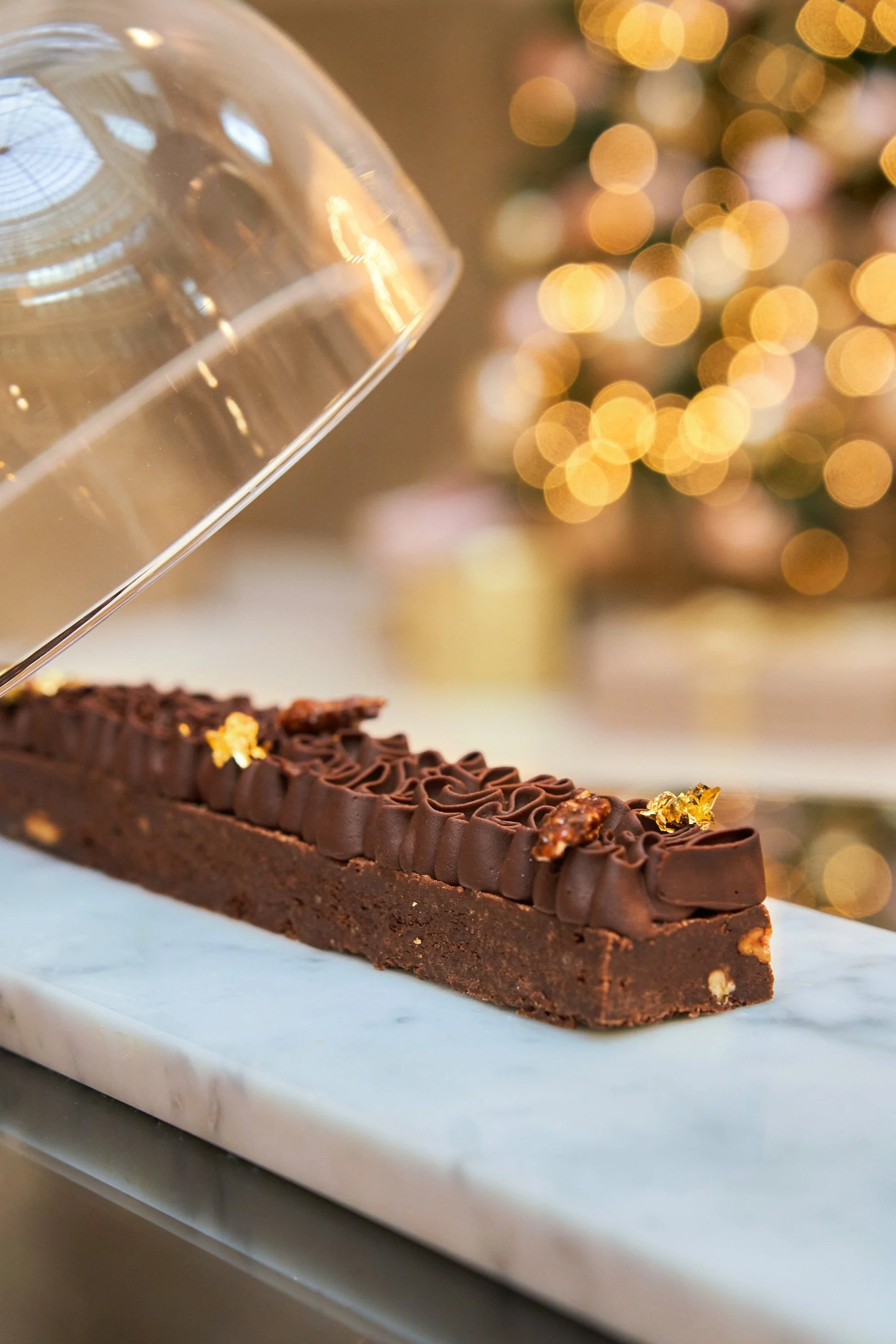 Une tarte au chocolat décorée de garnitures en or comestible, sur une plaque en marbre, avec un fond flou de lumières de Noël.