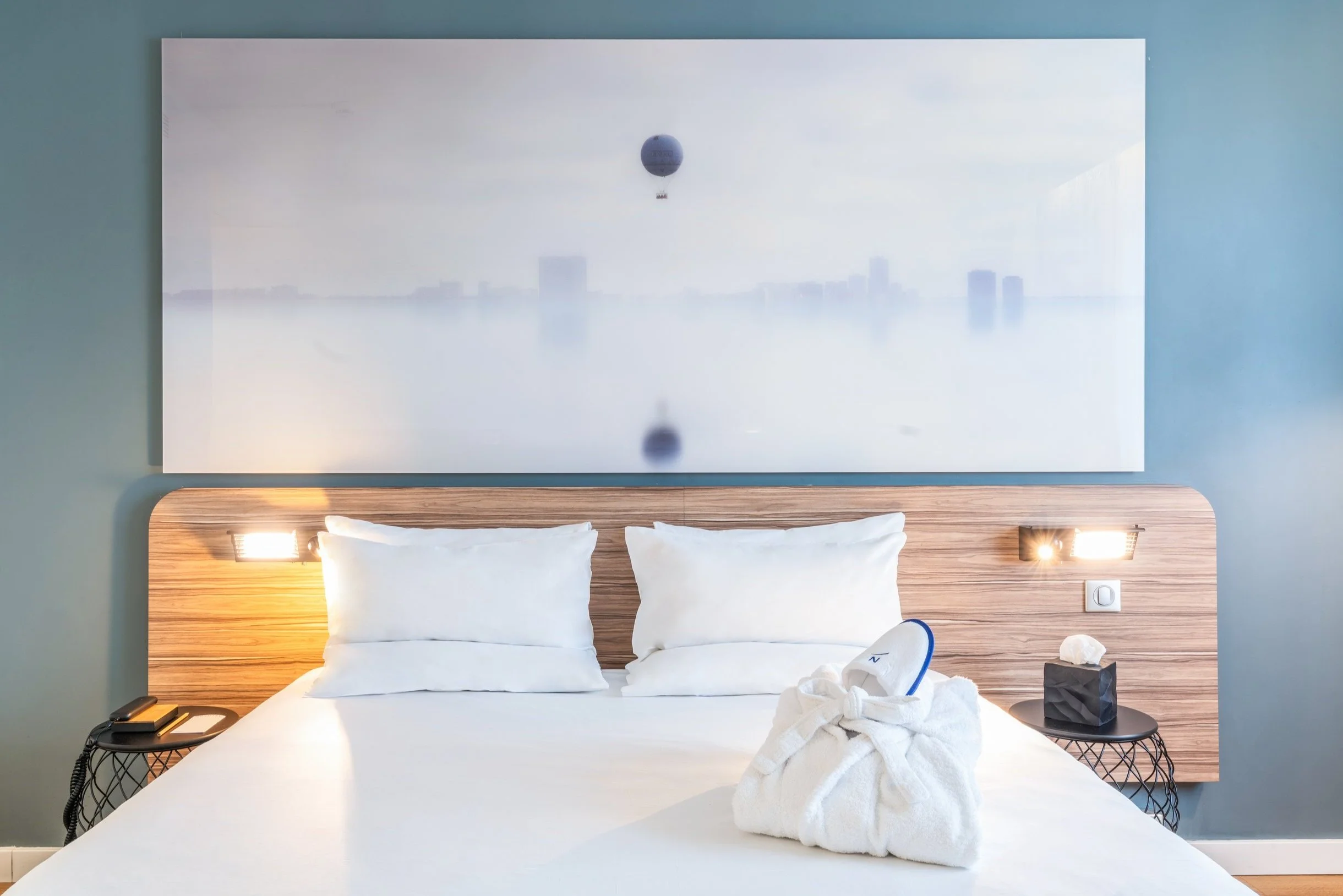 Lits dans une chambre d'hôtel avec une tête de lit en bois, deux lampes de lecture, un téléphone, une boîte de mouchoirs, un fer à repasser dans un drap, et un tableau au-dessus du lit représentant un paysage avec un ballon à air chaud.
