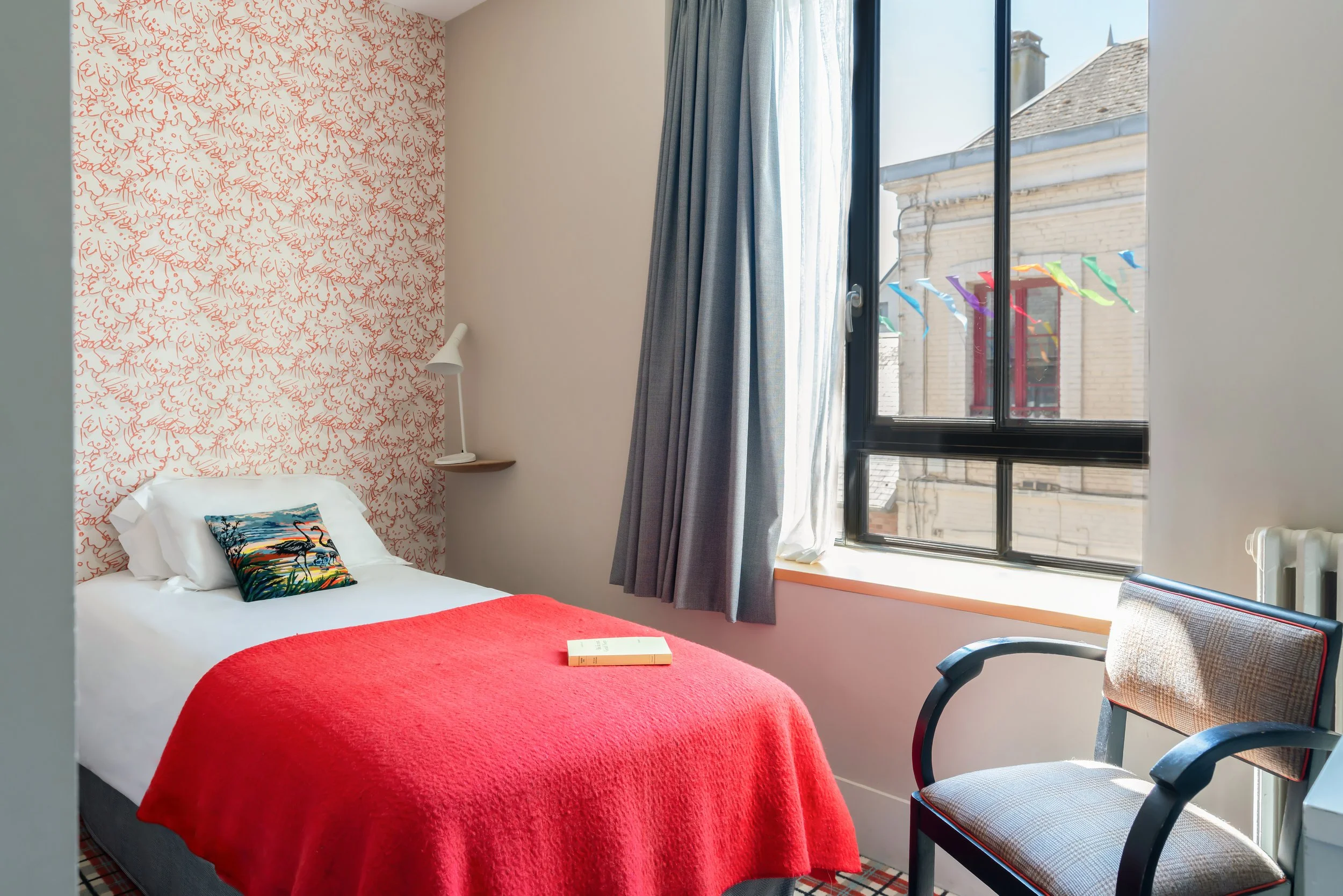 Chambre avec un lit simple recouvert d'une couverture rouge, une bibliothèque, un fauteuil, un mur avec papier peint rouge et blanc, une grande fenêtre avec rideaux gris et un décor coloré à l'extérieur.