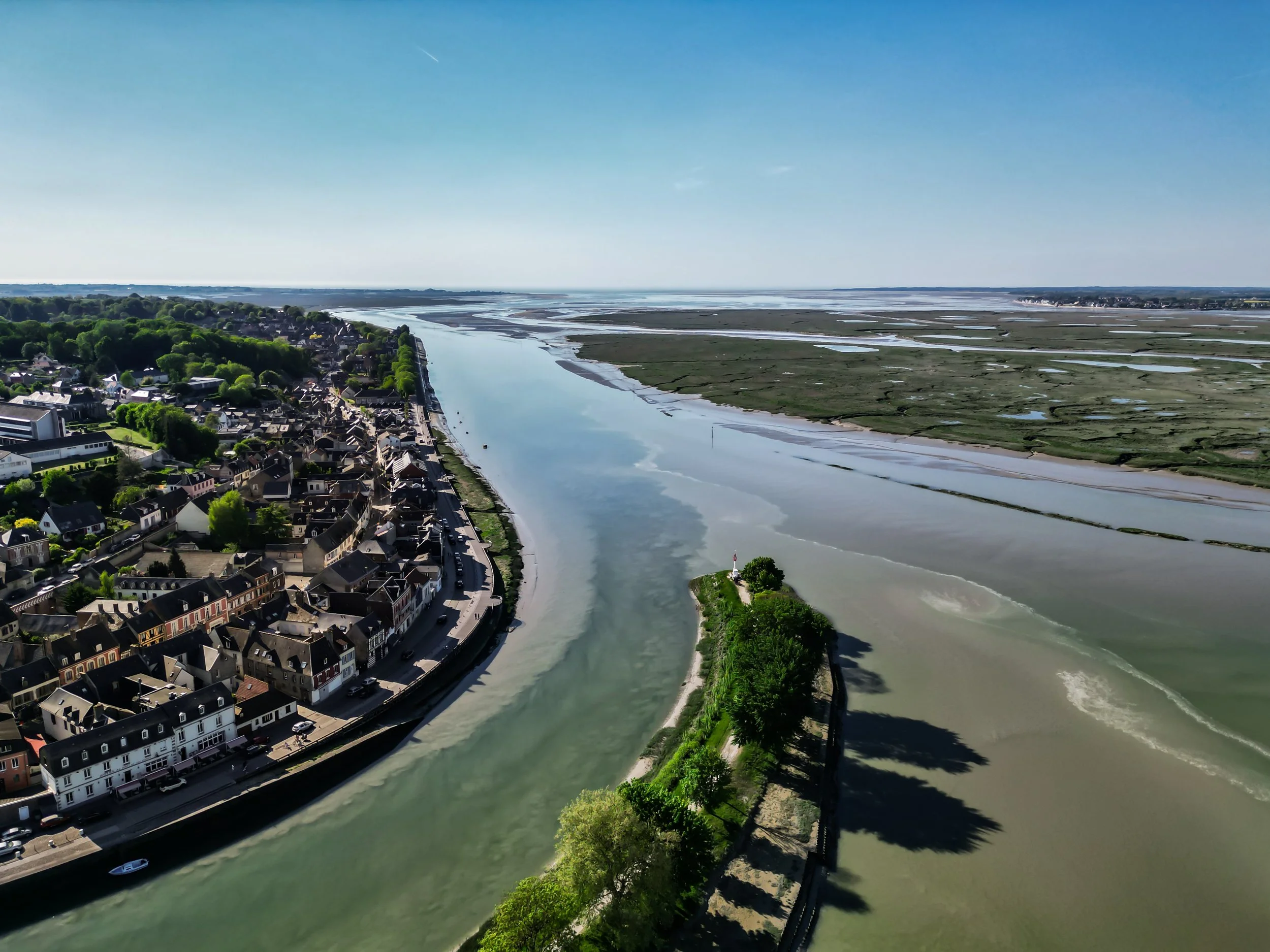 Vue aérienne d'une ville bordant une rivière qui se divise en plusieurs bras, avec une zone marécageuse en arrière-plan, ciel bleu clair.