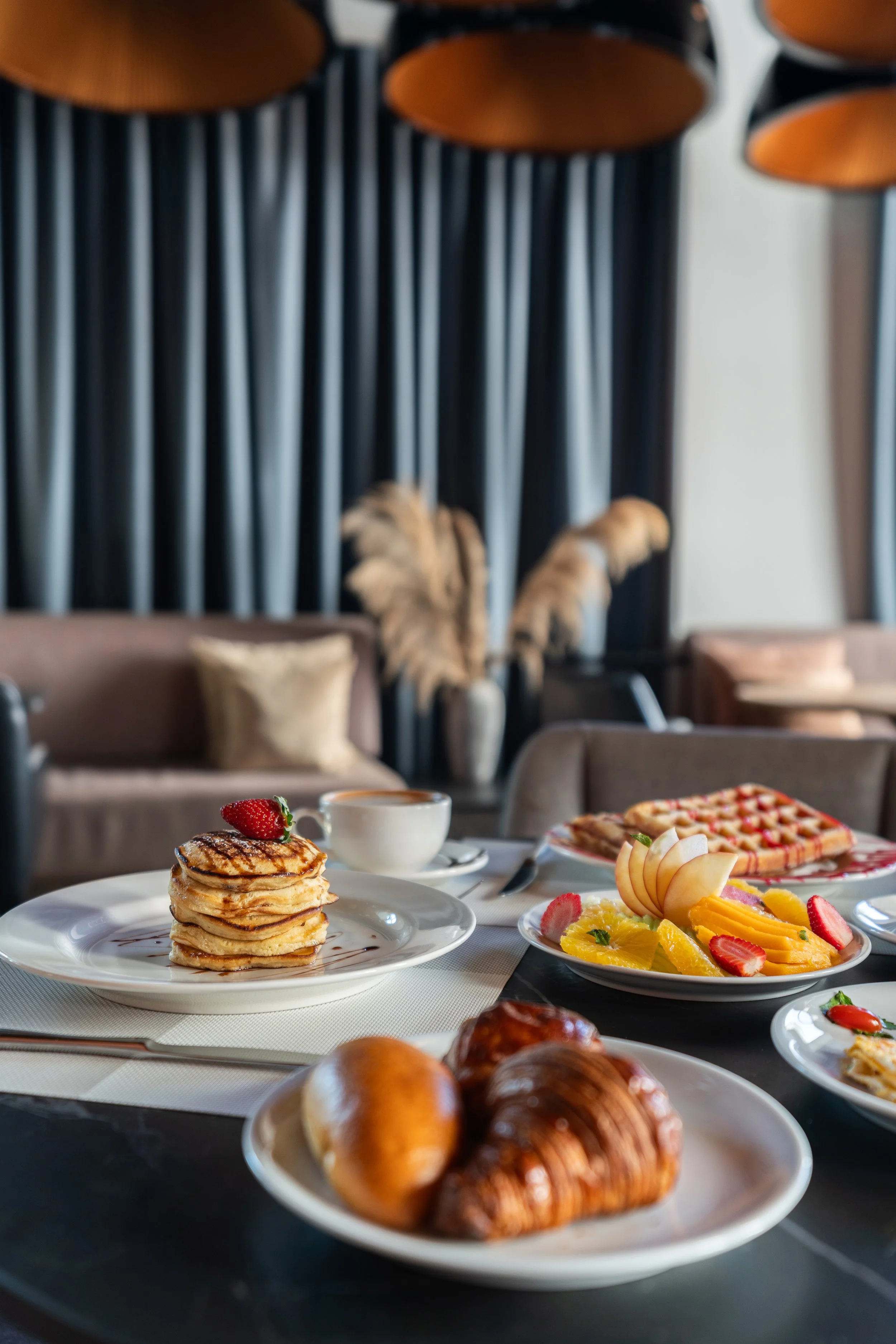 Petit-déjeuner avec des pancakes garnis de fraises, un cappuccino, une assiette de fruits frais, des croissants et des pâtisseries dans un cadre élégant