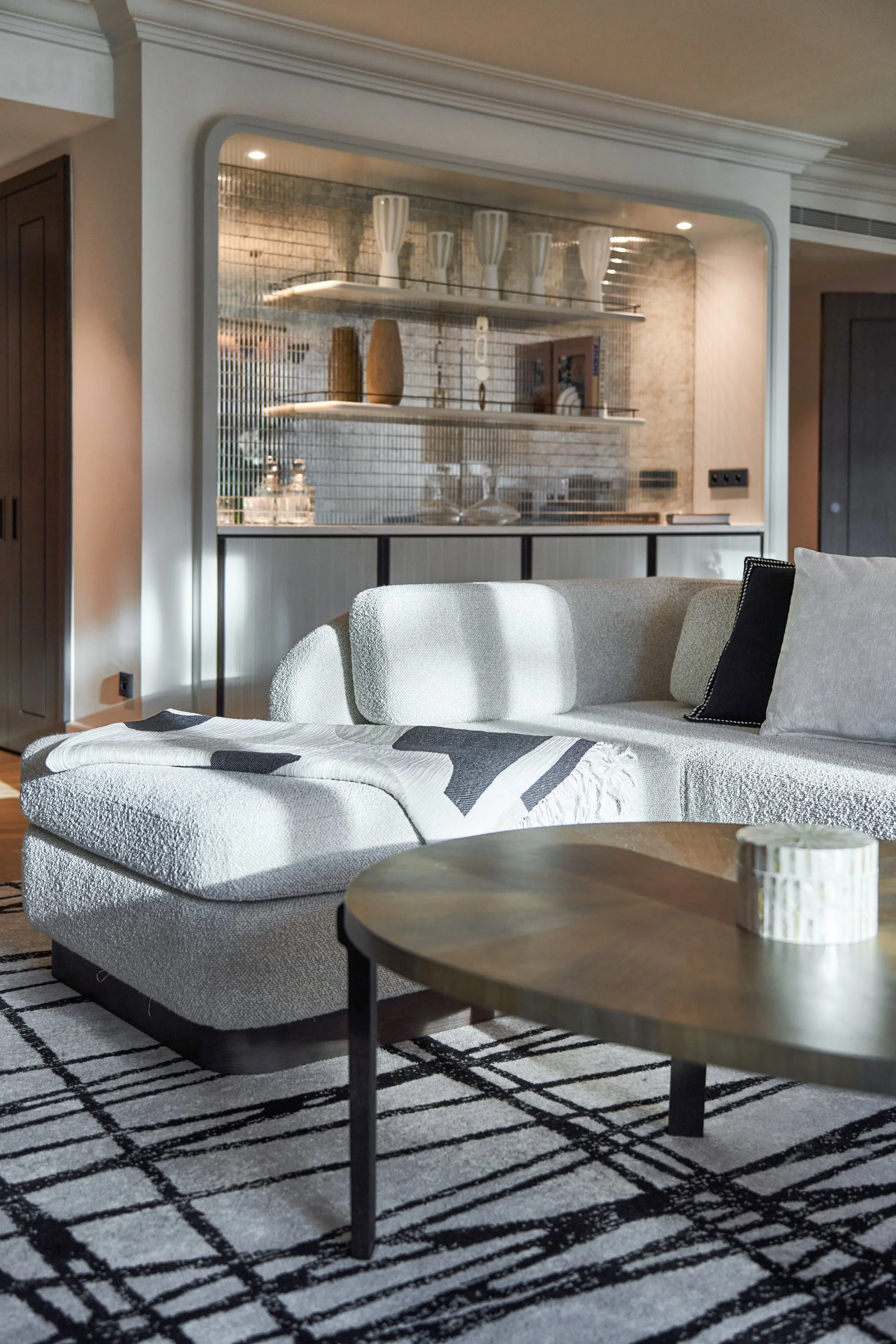 Intérieur d’un salon moderne avec canapé beige, table basse en bois, tapis noir et blanc, et étagère en arrière-plan avec des vases blancs et des objets décoratifs.