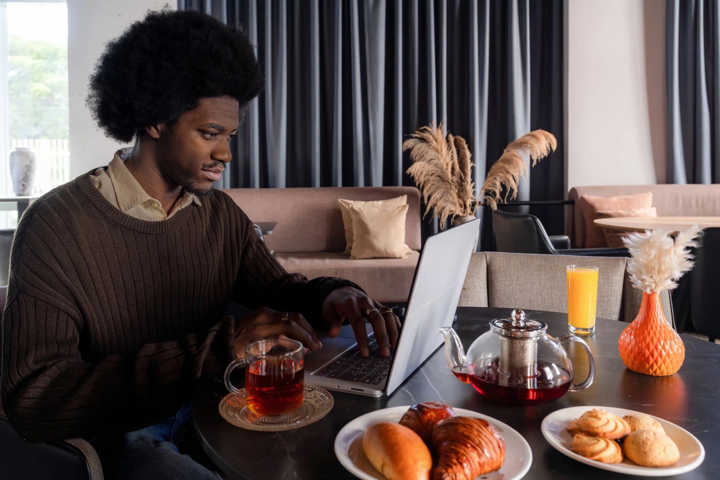 Homme utilisant un ordinateur portable lors d'un petit déjeuner avec café, thé, jus d'orange et viennoiseries dans un intérieur moderne et décoré de fleurs et coussins.