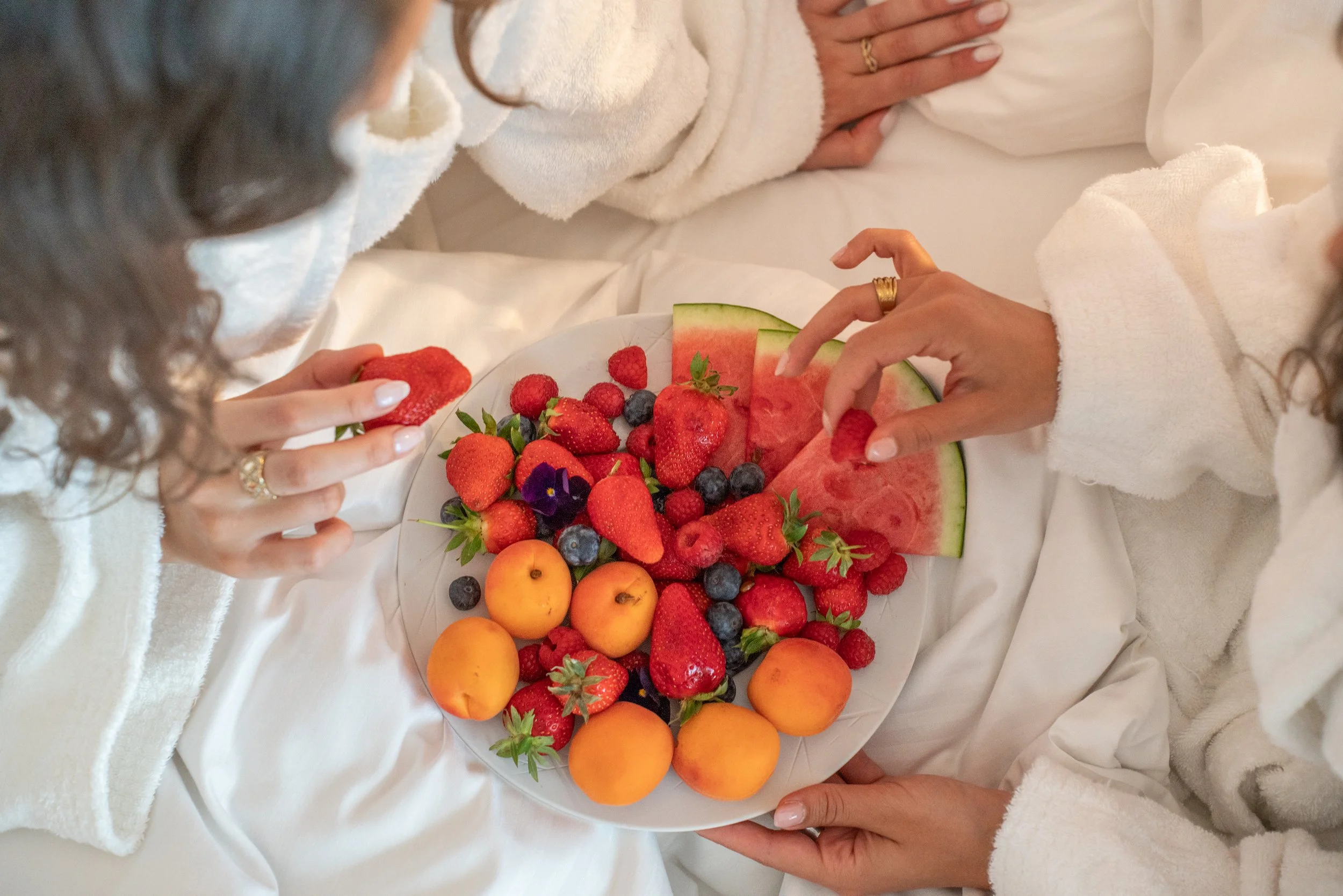 Deux femmes en peignoirs blancs partageant un plateau de fruits frais dont des fraises, pastèques, abricots, mûres, myrtilles et un violas violets.