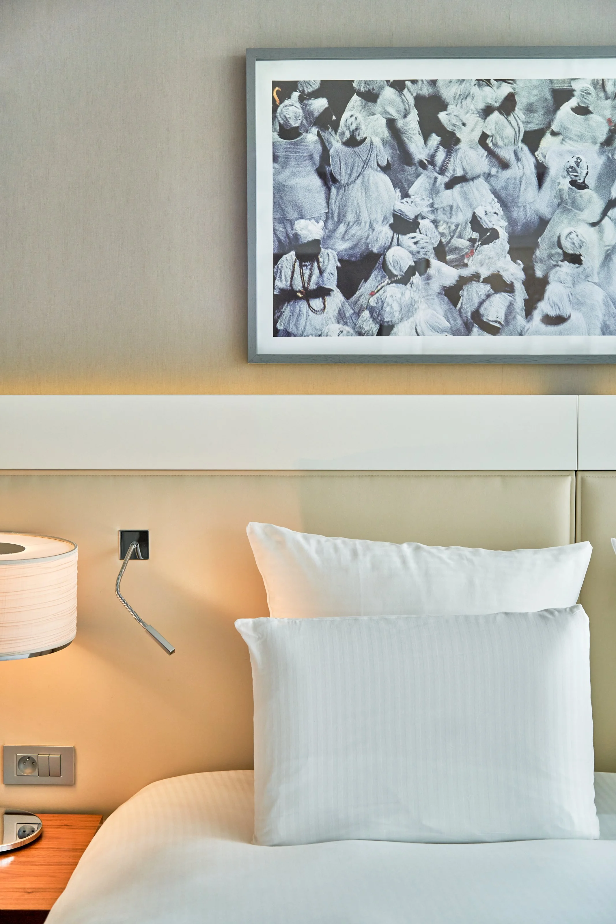 Chambre d'hôtel avec lit fait, deux oreillers, une lampe de table, une photo en noir et blanc d'une foule portant des vêtements traditionnels au mur.