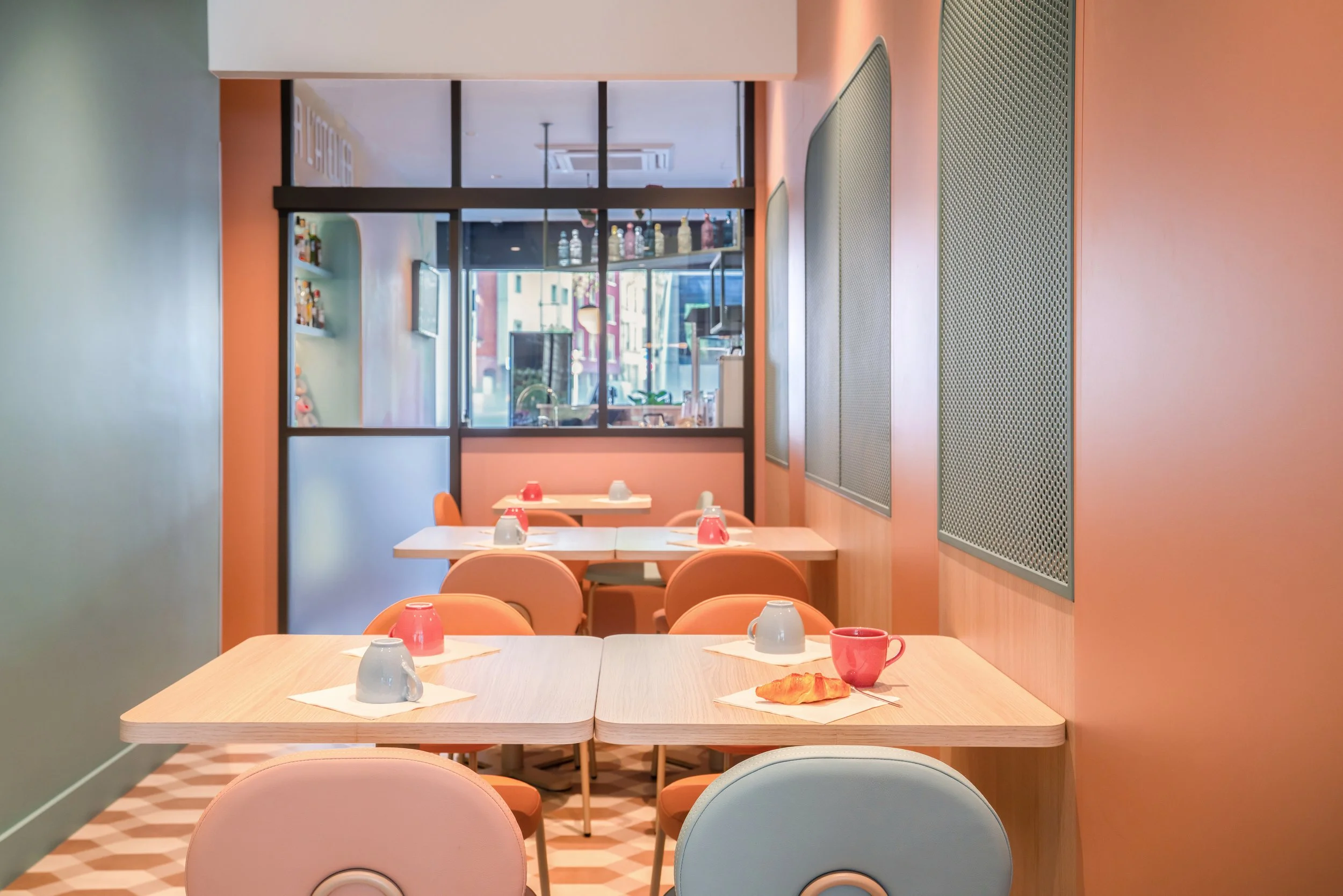 Salle de petit déjeuner coloré avec tables et tasses, mur orange, et vue vers la cuisine ou comptoir à travers une fenêtre en verre