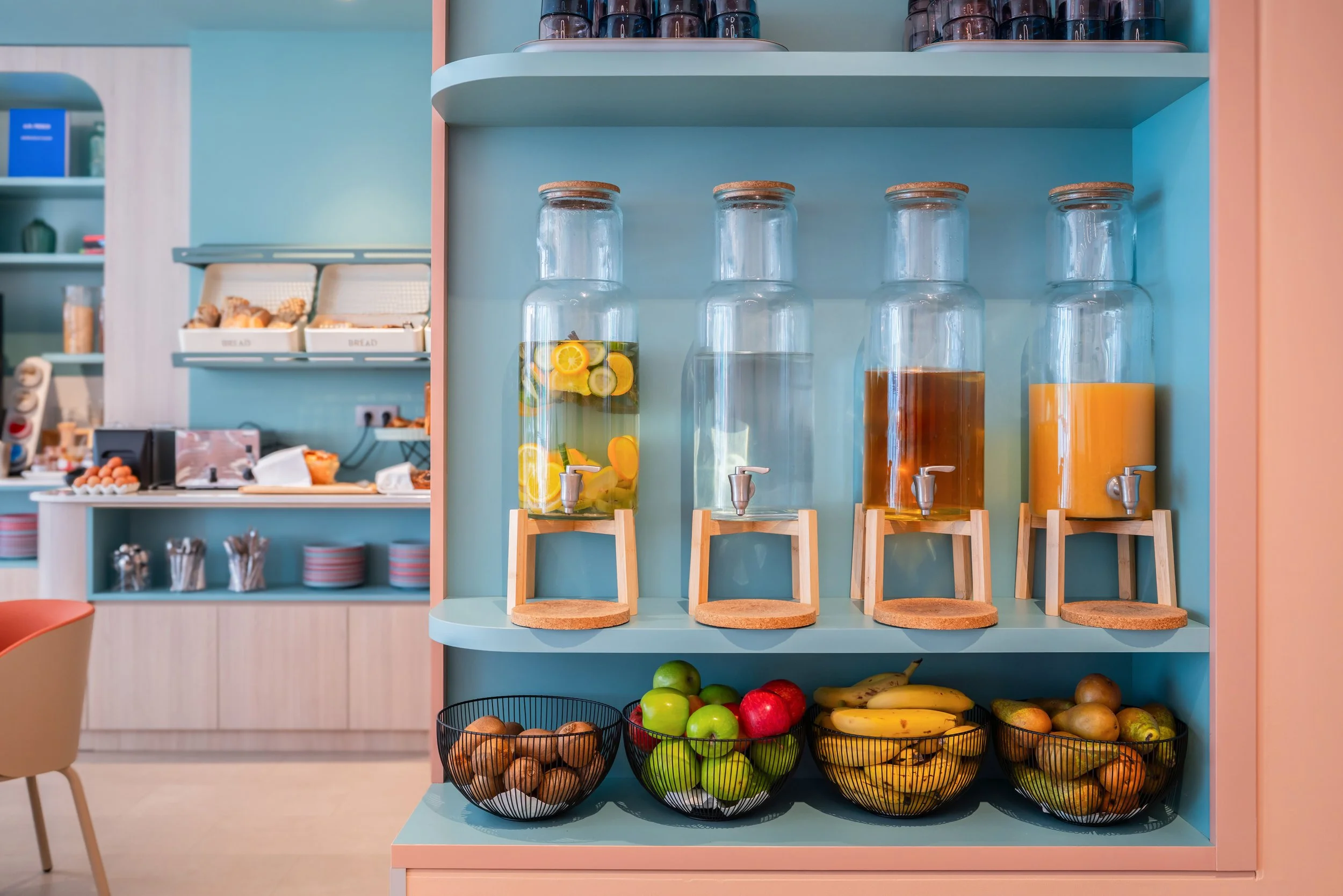 Distributeur de boissons avec des jarres en verre contenant de l’eau aromatisée à des fruits, fruits frais dans des paniers en fil métallique, dans un espace de buffet ou de cafétéria coloré.