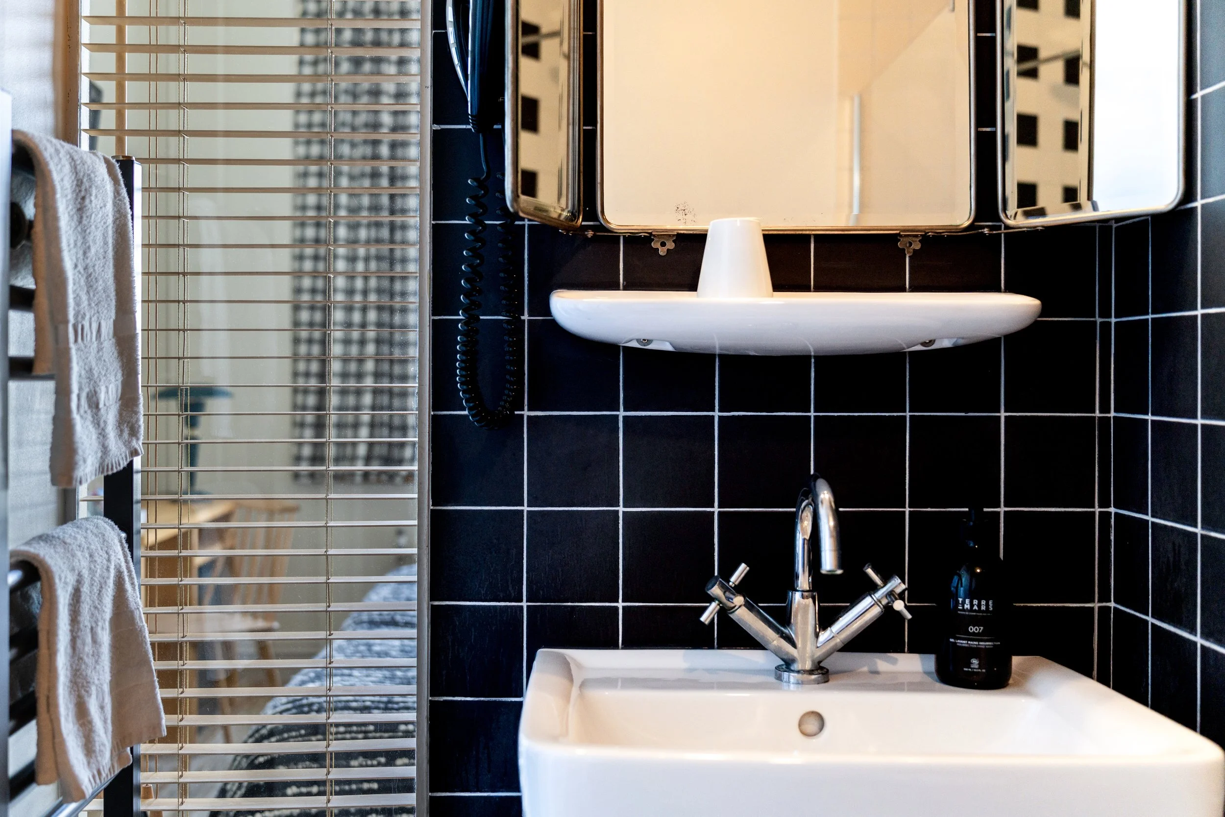 Lavabo blanc avec robinet en métal, miroir au-dessus, bouteille de parfum noire à côté, murs en carreaux noirs, serviettes blanches sur un support métallique, et un sèche-linge ou radiateur blanc avec grille à gauche.