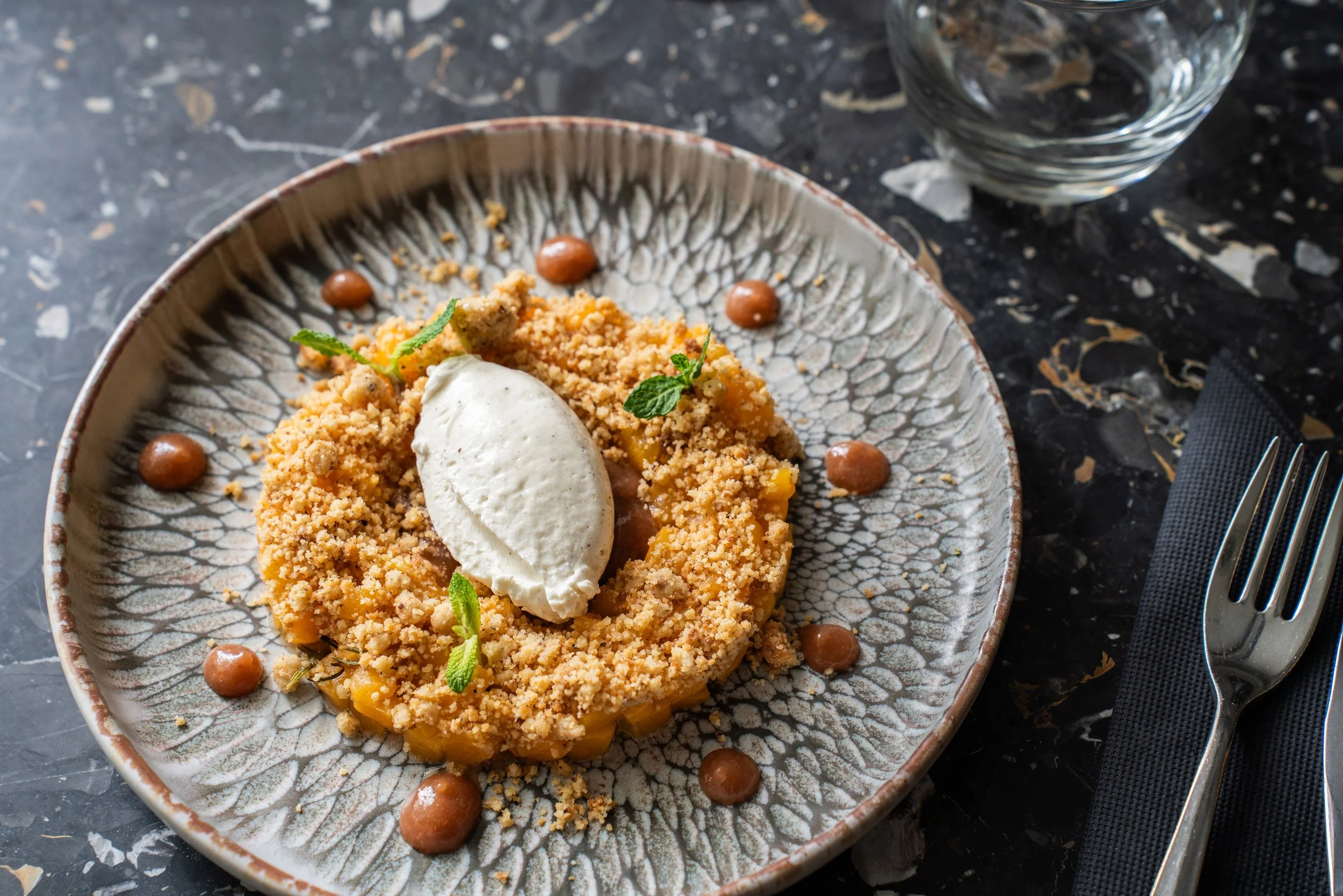 Crumble, crème fouettée, menthe et pépites de chocolat sur une assiette en céramique. Également une verre d'eau à côté sur une table en marbre noir.