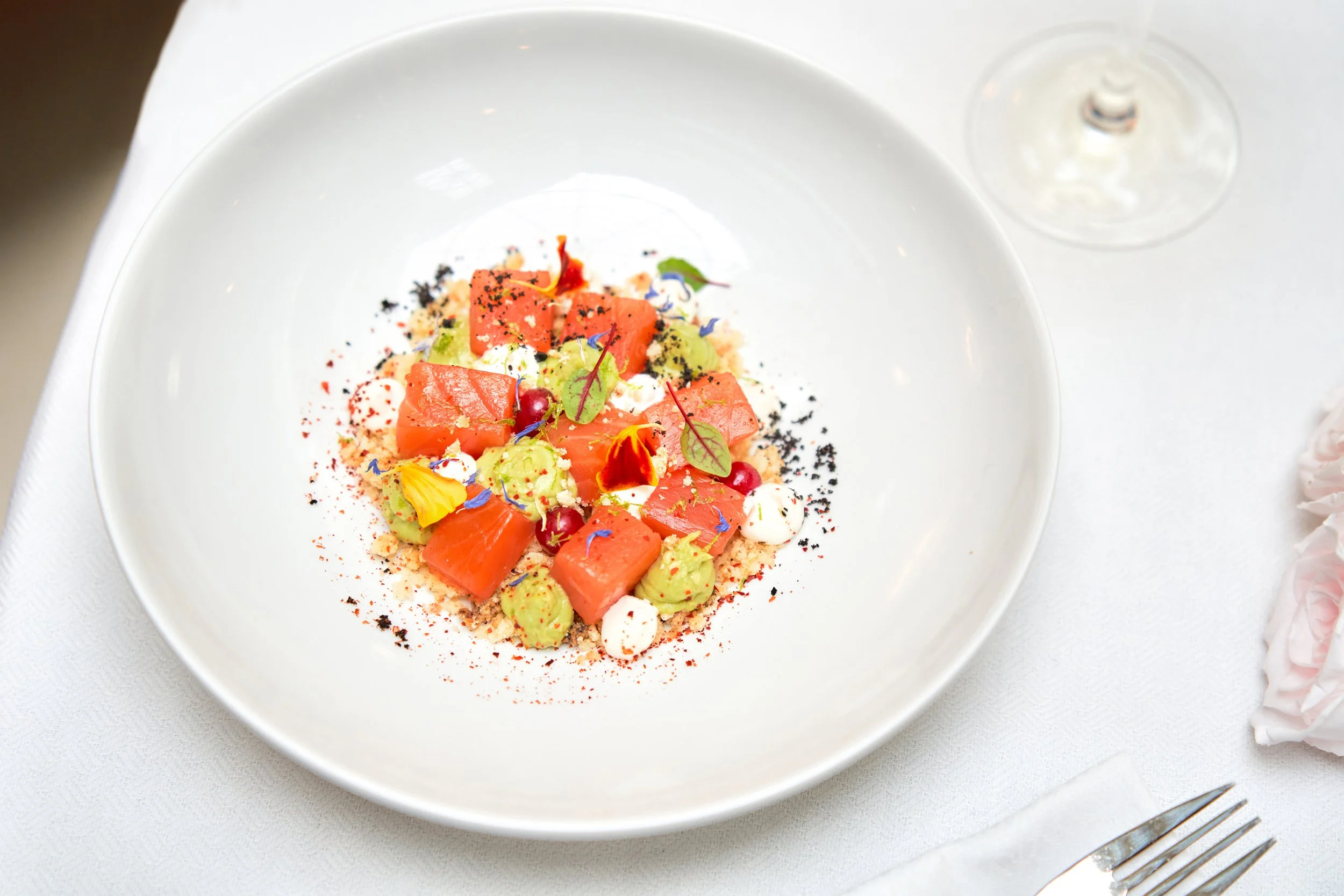 Tartare de saumon, avocat, graines, micro-pousses et fleurs comestibles dans une assiette blanche.
