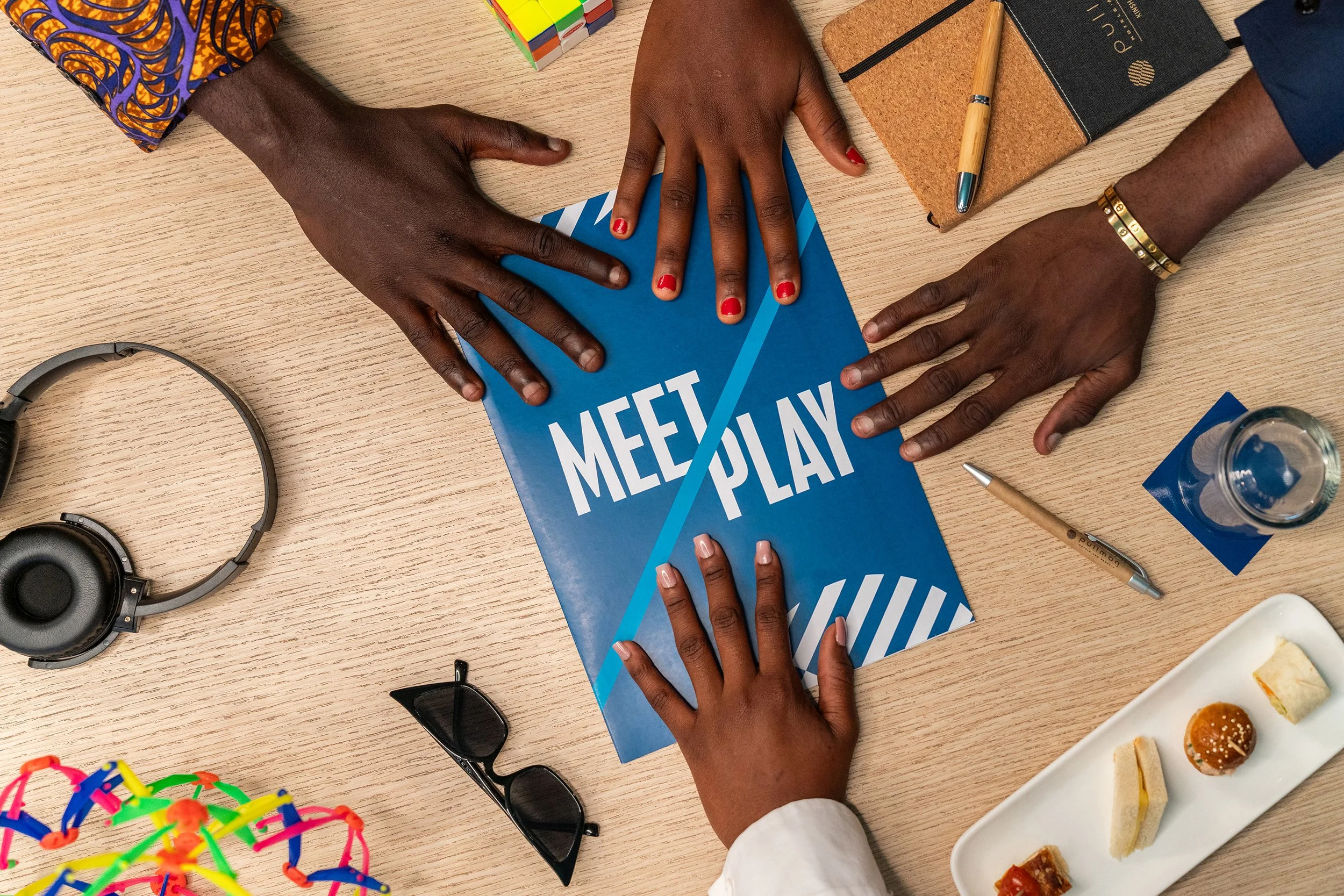 Des personnes posant leur main sur une bloc note 'Meet & Play' sur une table en bois, entourée d'accessoires de bureau, des lunettes de soleil, une paire d'écouteurs, un stylo, une bouteille d'eau, et une assiette avec des petits gâteaux.