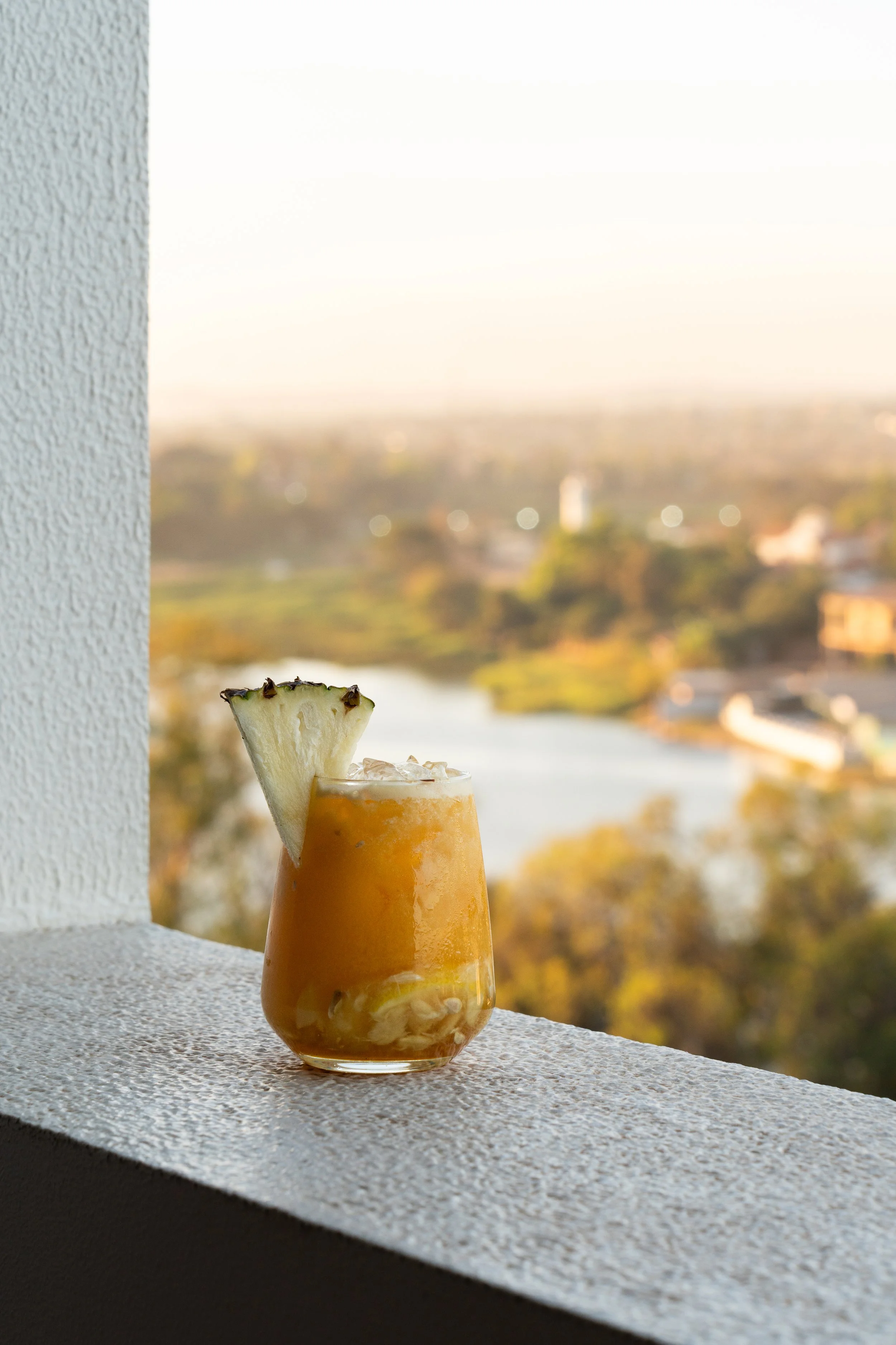 Verre de cocktail orange avec une tranche d'ananas en décoration, placé sur un rebord de fenêtre avec un paysage flou en arrière-plan