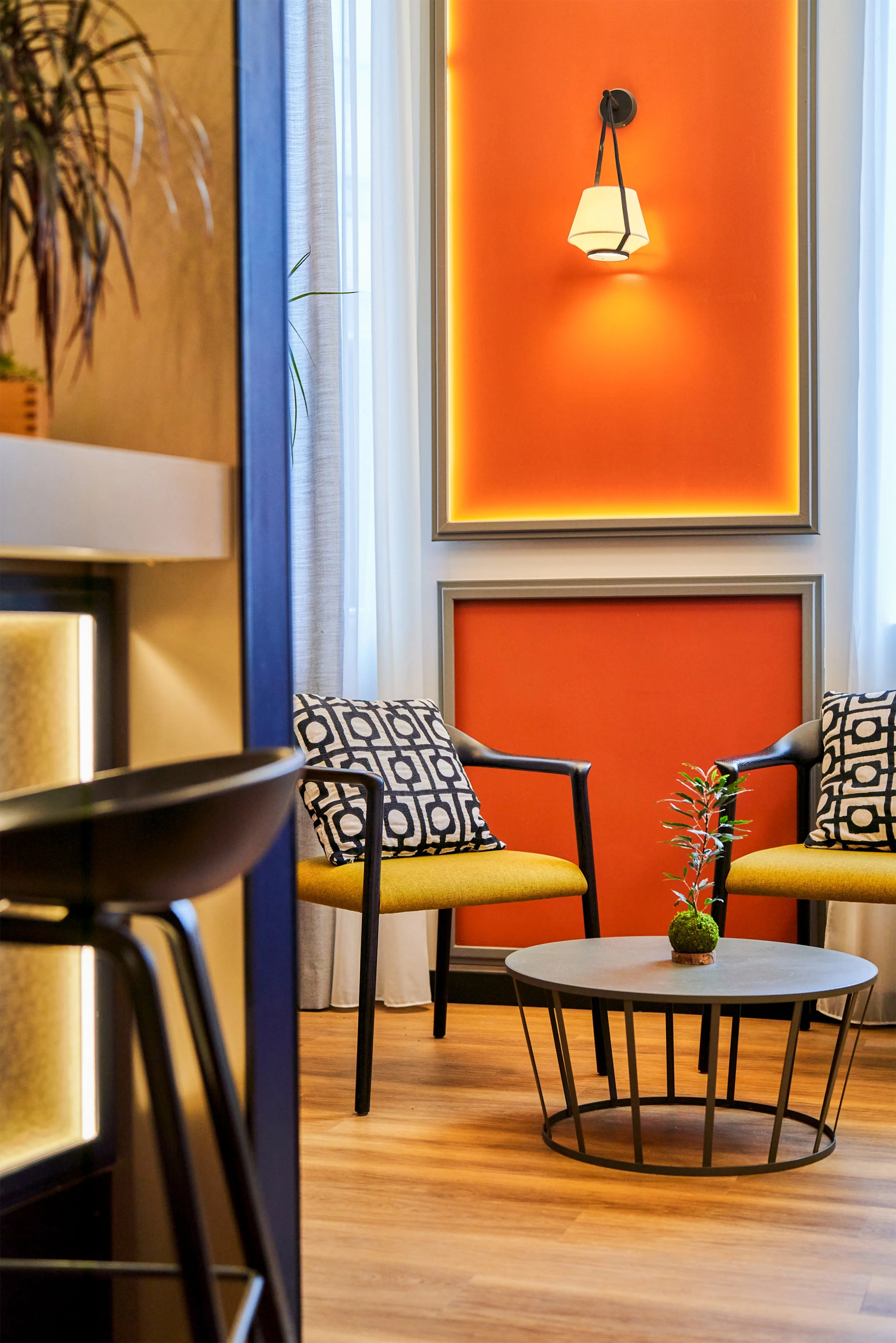 Coin salon avec deux fauteuils à coussins géométriques, table basse ronde avec un petit pot de plantes, mur orange avec éclairage intégré et lampe suspendue, rideaux blancs et sol en bois.