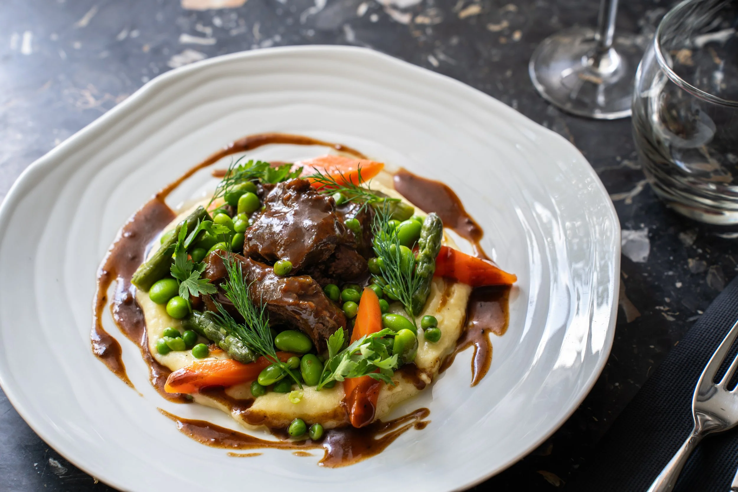 Plat de viande en sauce avec purée, carottes, haricots verts et herbes fraîches, présenté sur une assiette blanche
