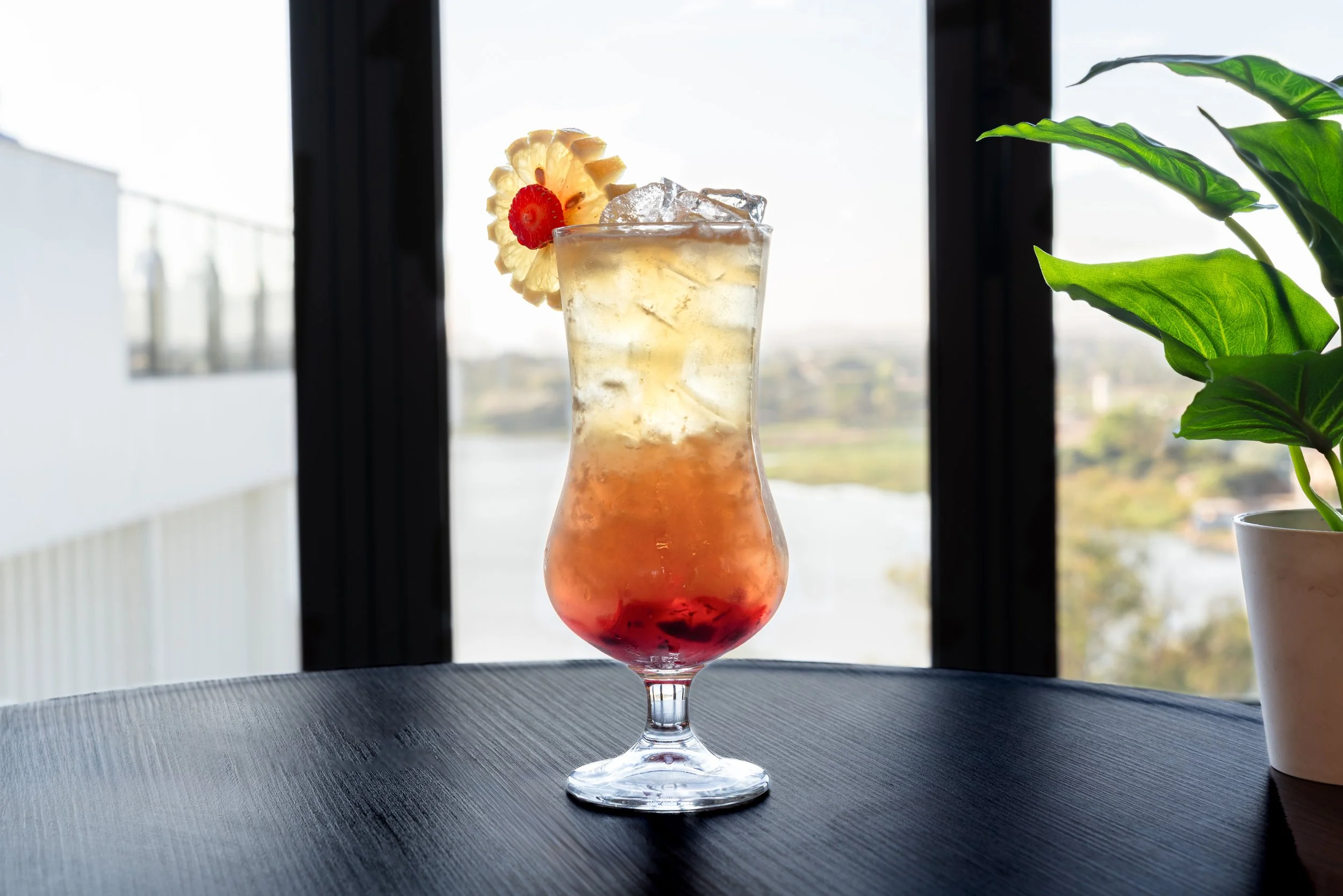 Cocktail coloré dans un verre tulipe avec une tranche d'ananas et une cerise, placé sur une table noire avec une plante verte à droite, devant une fenêtre avec vue sur l'extérieur