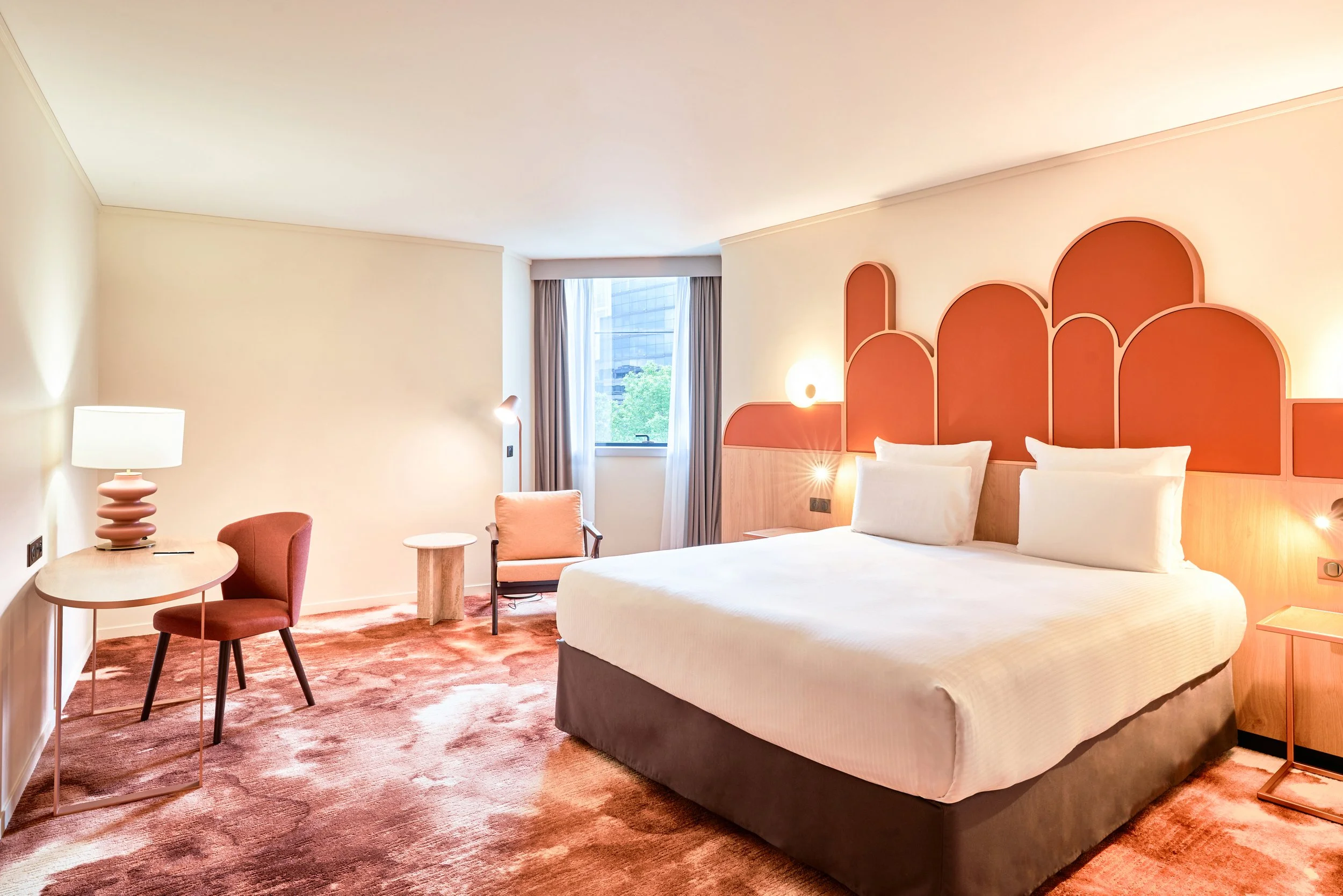 Chambre d'hôtel moderne avec un lit king-size, un bureau avec lampe, fauteuils et tapis orange.