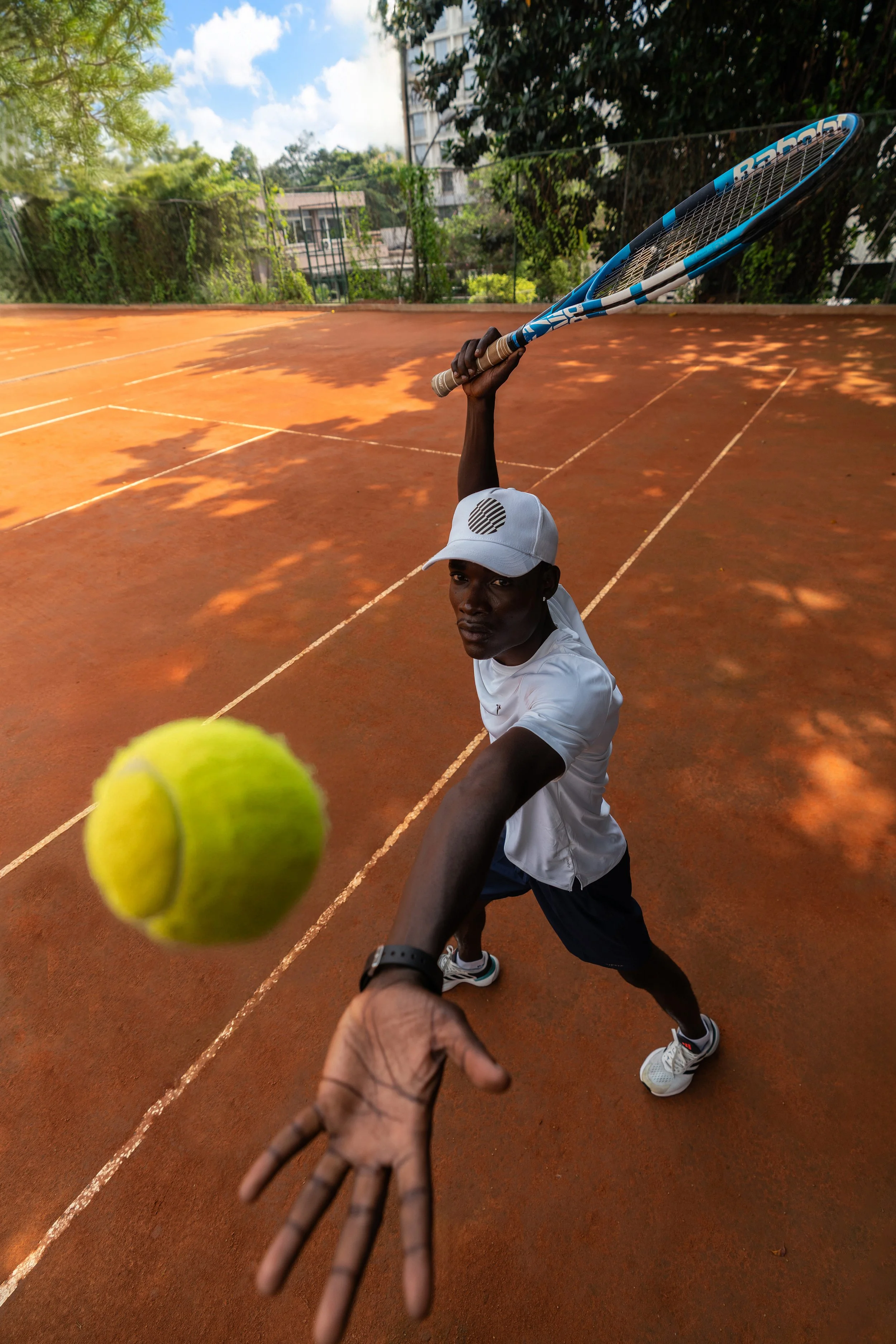 Un joueur de tennis noir en position de frappe, tenant une raquette bleue dans une main, une balle de tennis en gros plan dans l'objectif, sur un court en terre battue en extérieur.