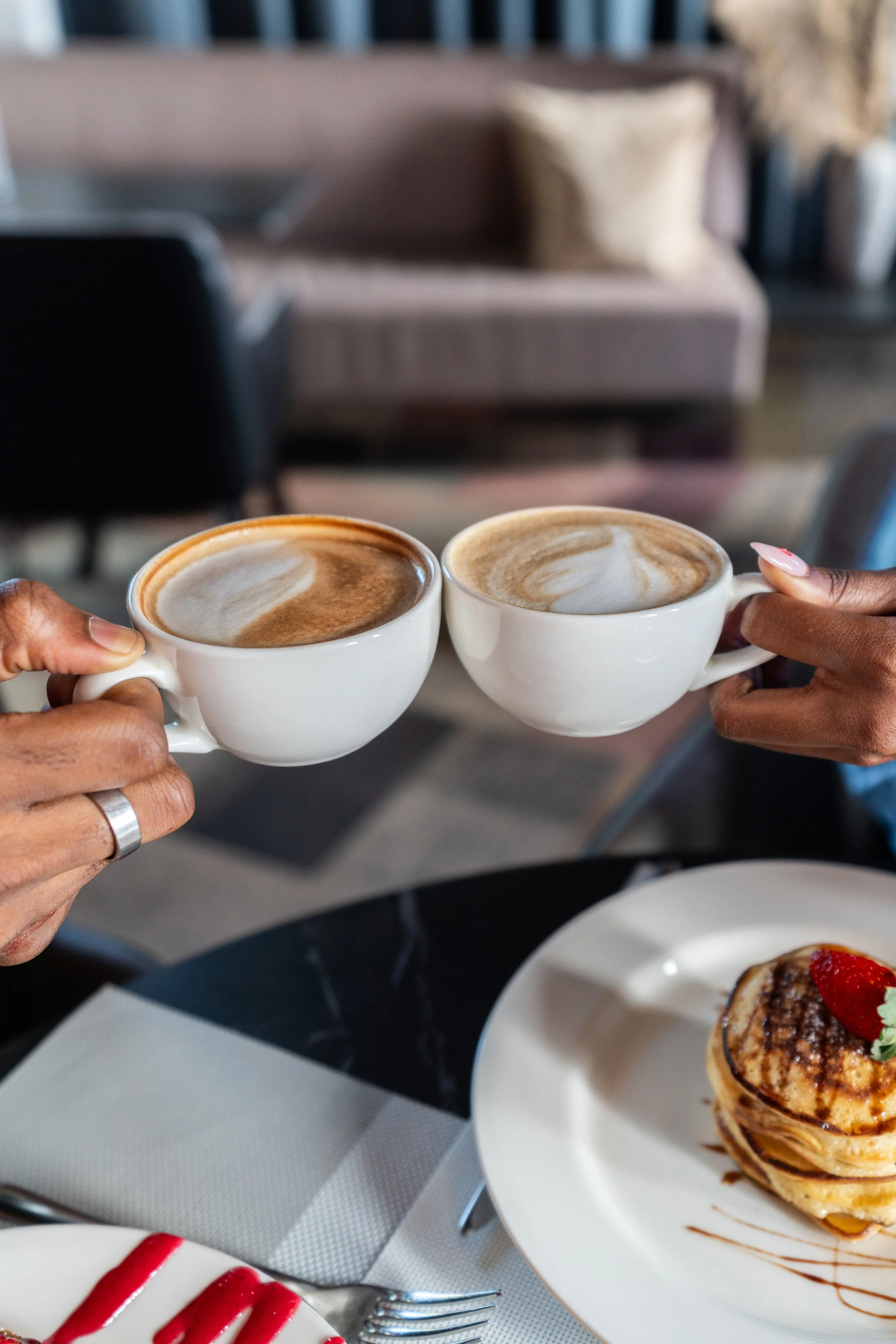 Deux personnes tenant des tasses de café latte avec art latte, à une table où se trouve aussi un dessert avec des fraises et du chocolat.