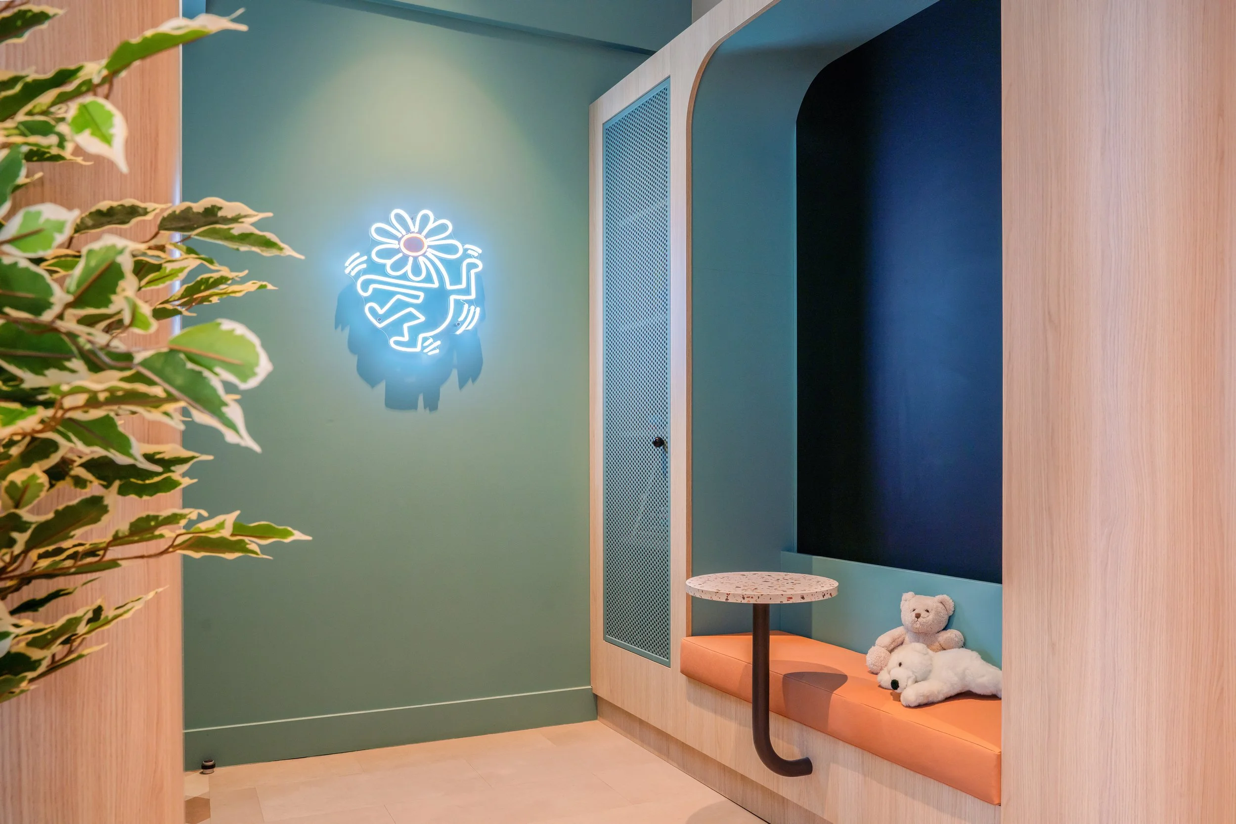 Coin salon avec un banc orange, deux ours en peluche, un petit tableau rond, un mur vert avec une enseigne lumineuse en forme de fleur avec un visage de smiley, plantes vertes à gauche, éléments en bois