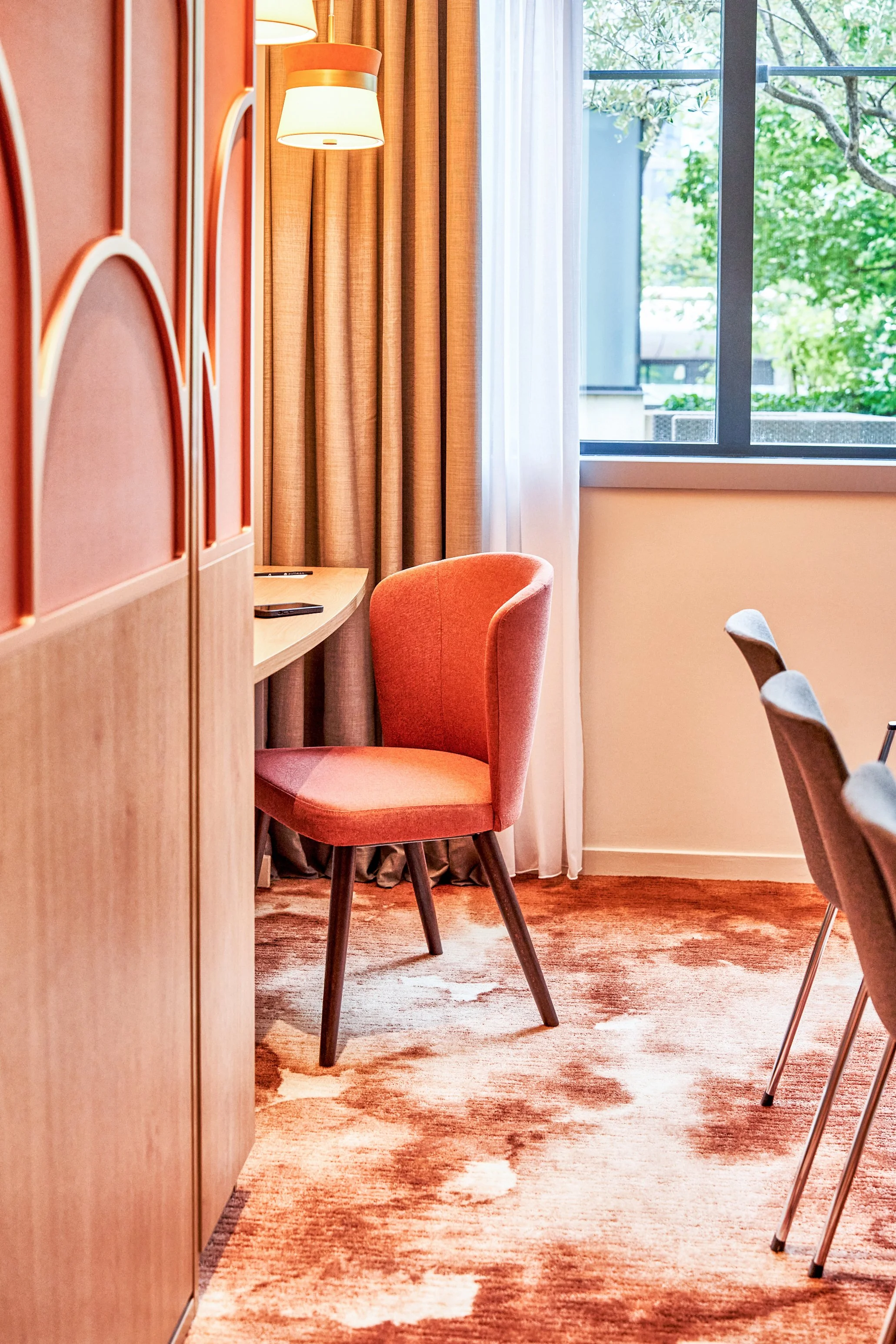 Coin de chambre avec fauteuil orange, grande fenêtre avec vue sur arbres, rideaux beige, lampe suspendue