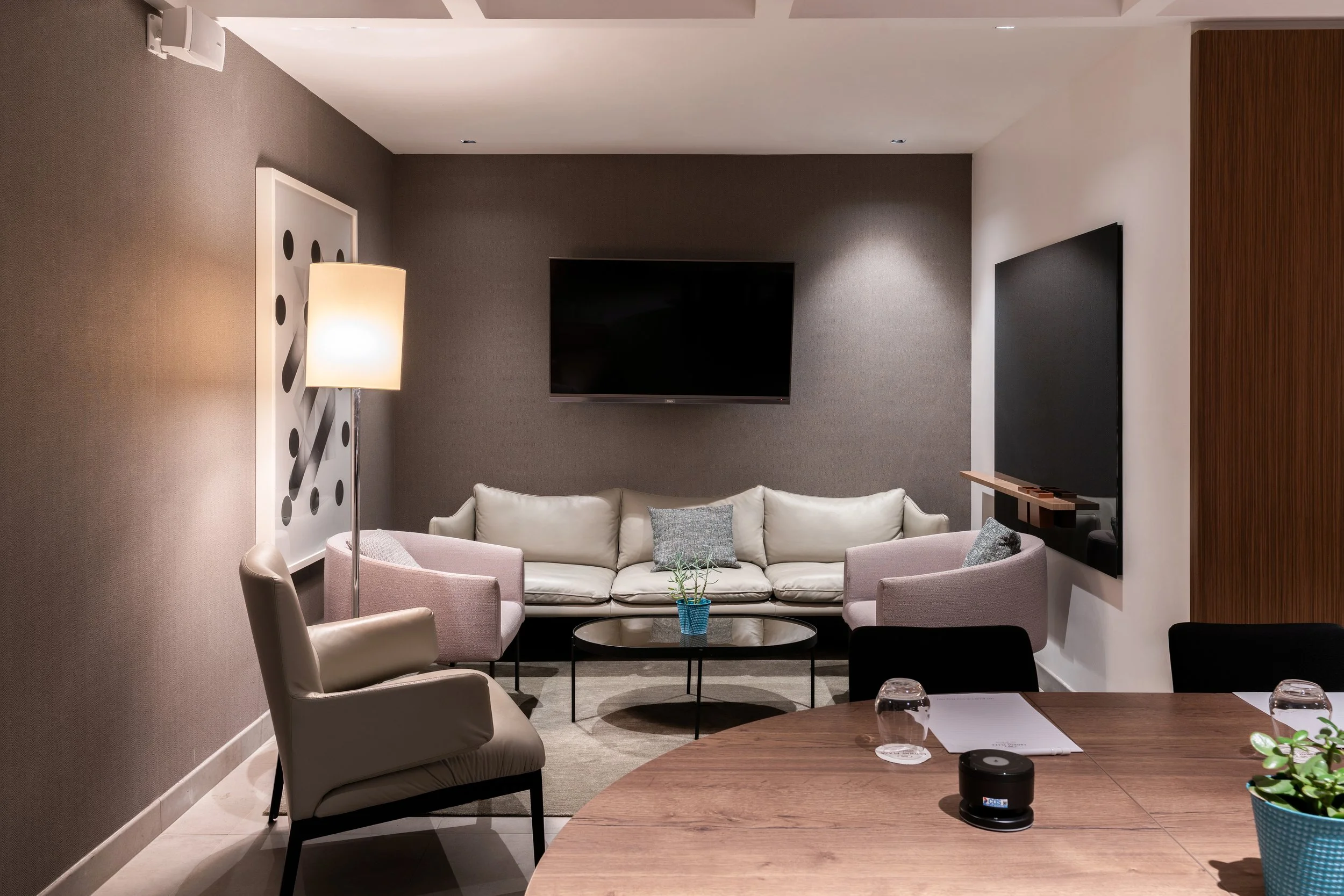 Salon moderne avec un canapé blanc, deux fauteuils roses, une table basse en verre avec une plante en pot turquoise, sur un fond de mur en deux tons avec téléviseur à écran plat, lampe sur pied et tableau décoratif.