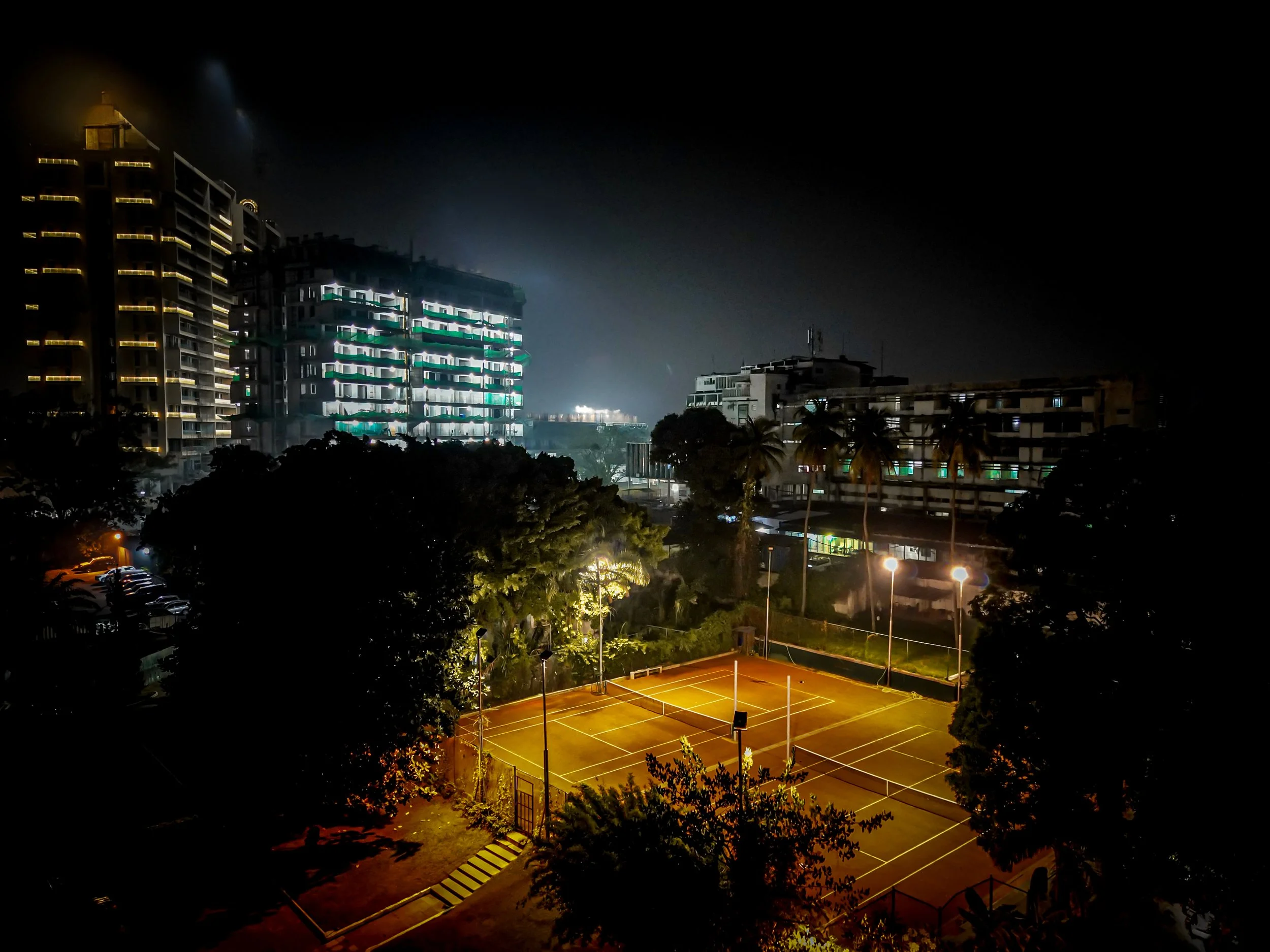 Terrains de tennis éclairés la nuit, entourés d'arbres et de bâtiments résidentiels modernes avec des lumières à l'étage.
