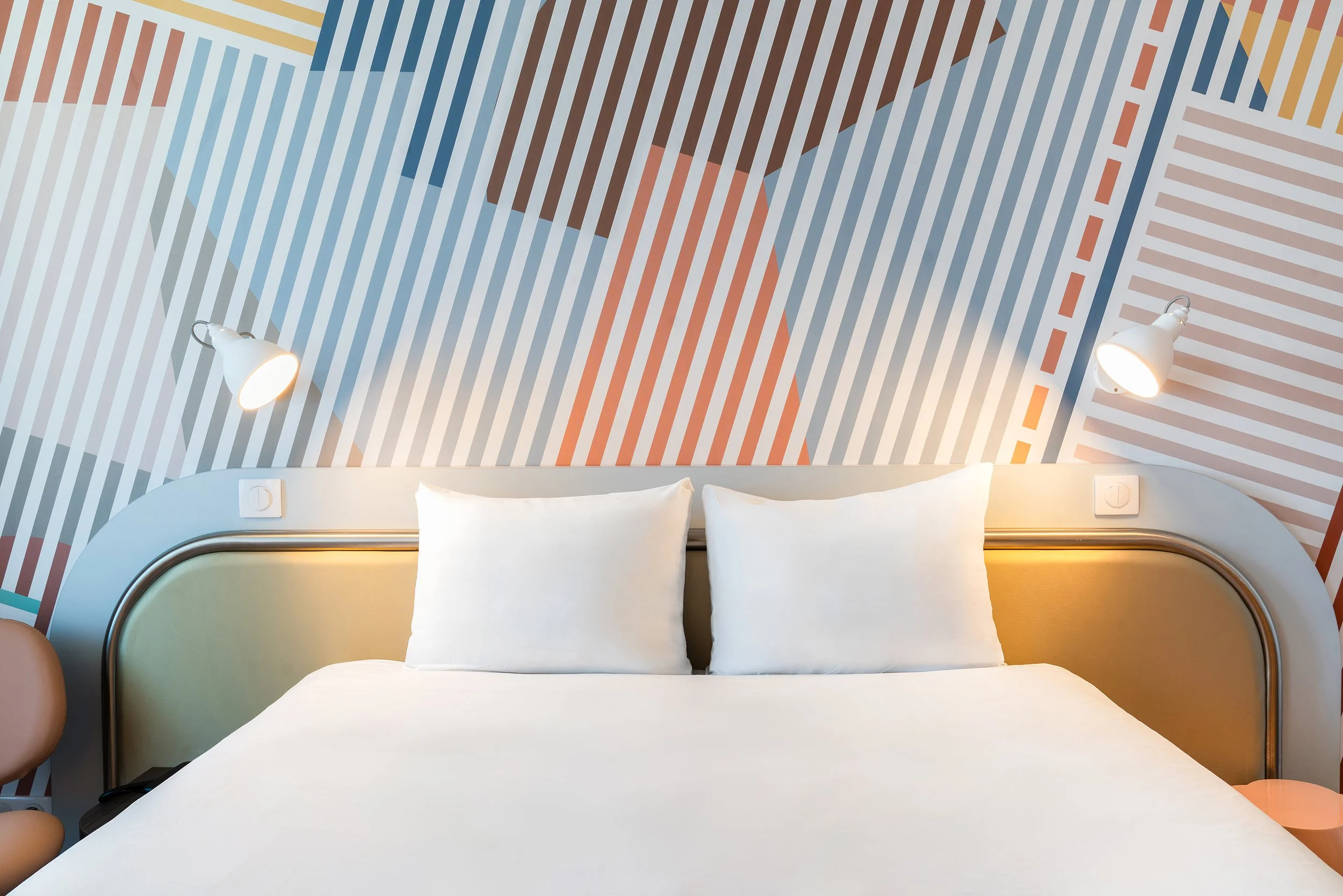 Chambre moderne avec un lit blanc à deux oreillers, décor mural abstrait avec lignes colorées et lampes de chaque côté de la tête de lit.