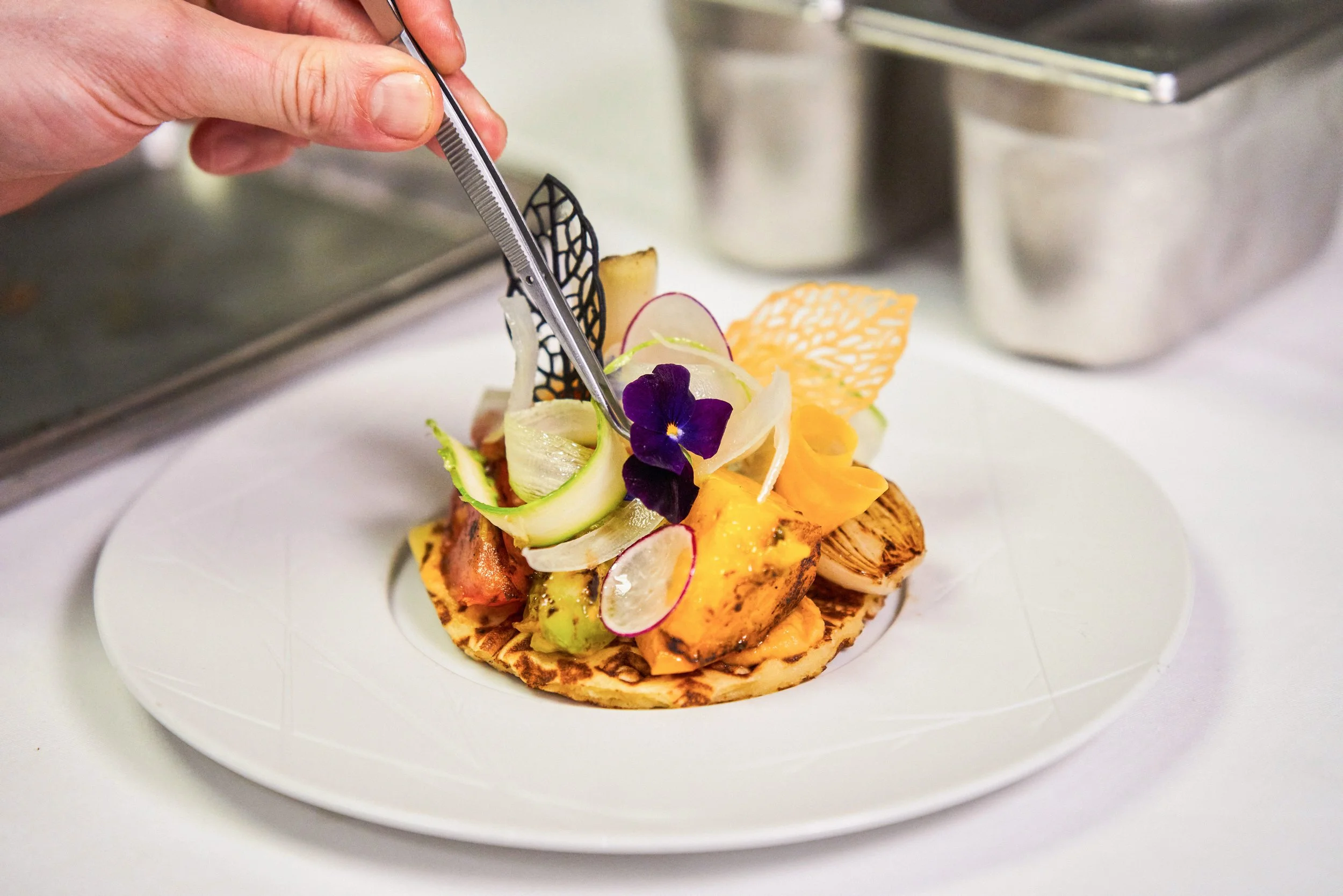 Chef décorant une assiette gastronomique avec des fleurs comestibles, des légumes et une présentation artistique sur une assiette blanche.