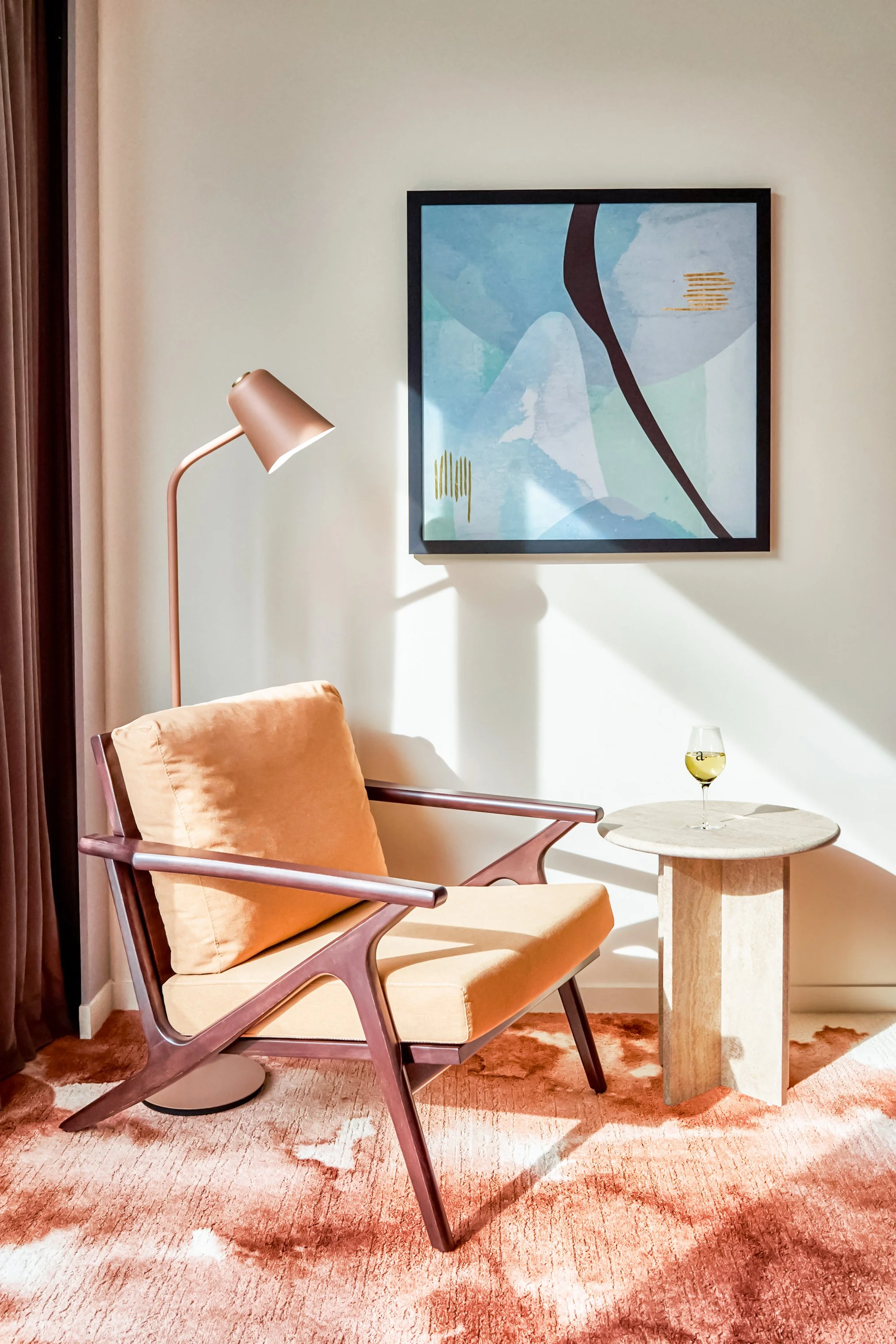 Coin salon avec un fauteuil en bois et tissu beige, une petite table ronde en pierre avec un verre de vin blanc, un lampadaire rose pâle, un tableau abstrait avec des formes dans des tons de bleu, vert et noir, et un tapis rose-orange.