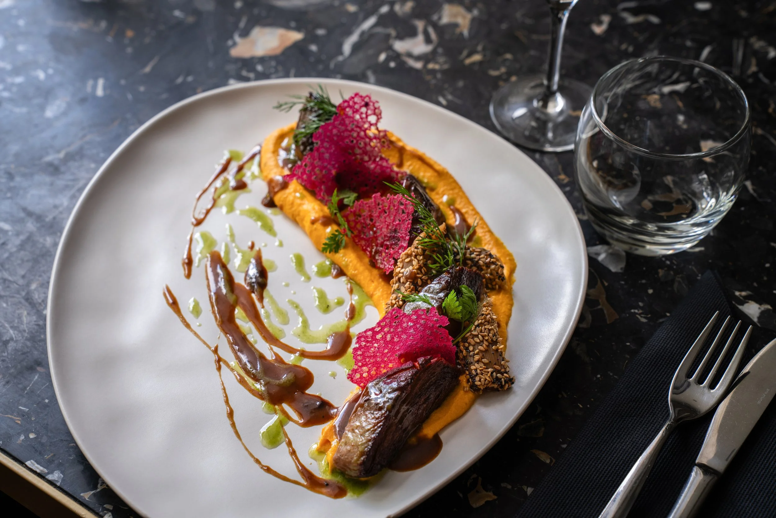 Assiette gastronomique avec un carré de viande, purée orange, garnitures croisées de croustillants rouges, graines de sésame, herbes fraîches, et sauces drizzled, à côté d'un verre d'eau et un verre de vin sur une table en marbre noir.