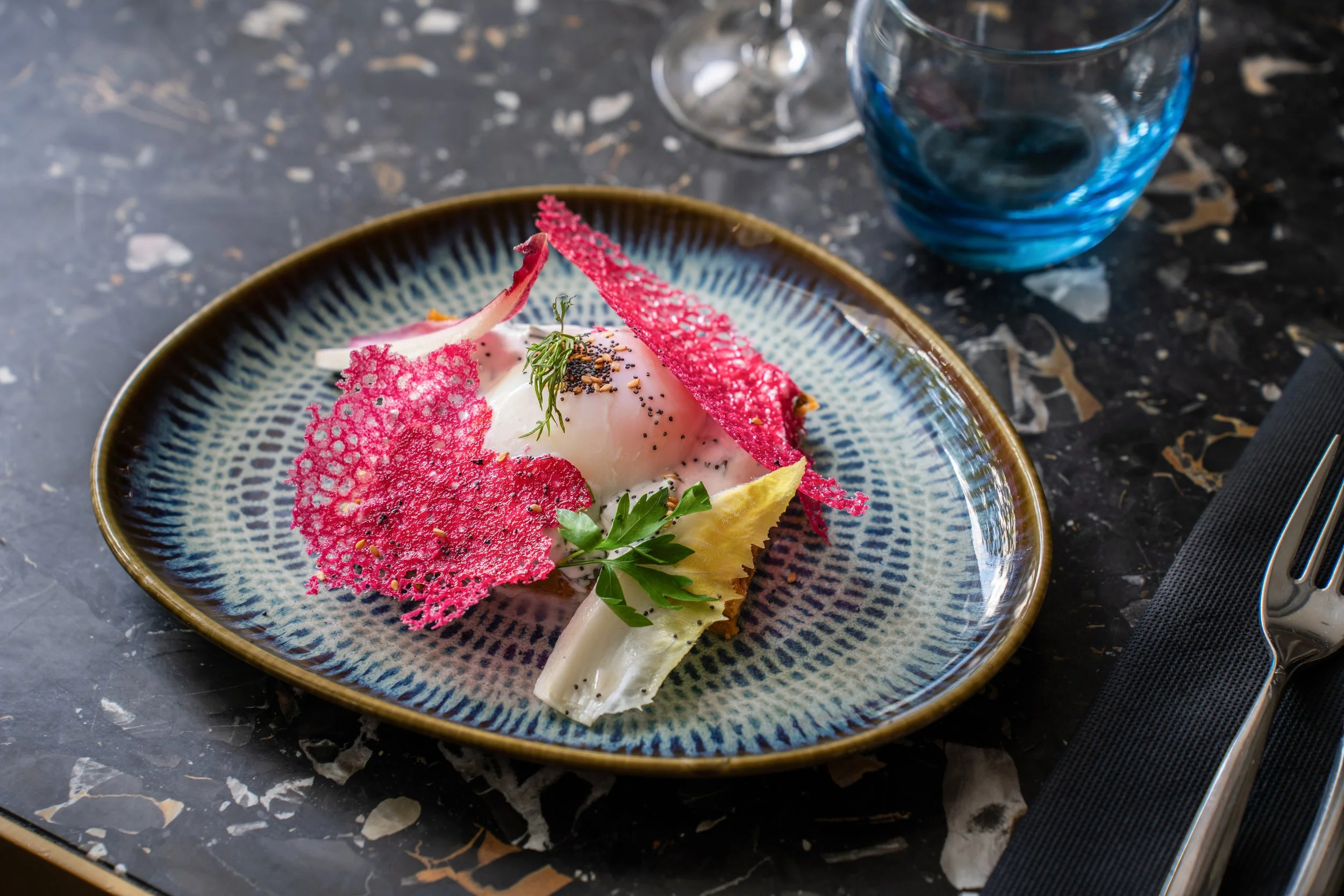 Assiette de présentation d'un plat gastronomique avec œuf poché, chips colorés et herbes, sur une table en marbre noir, accompagnée de verre d'eau et de verre de vin.