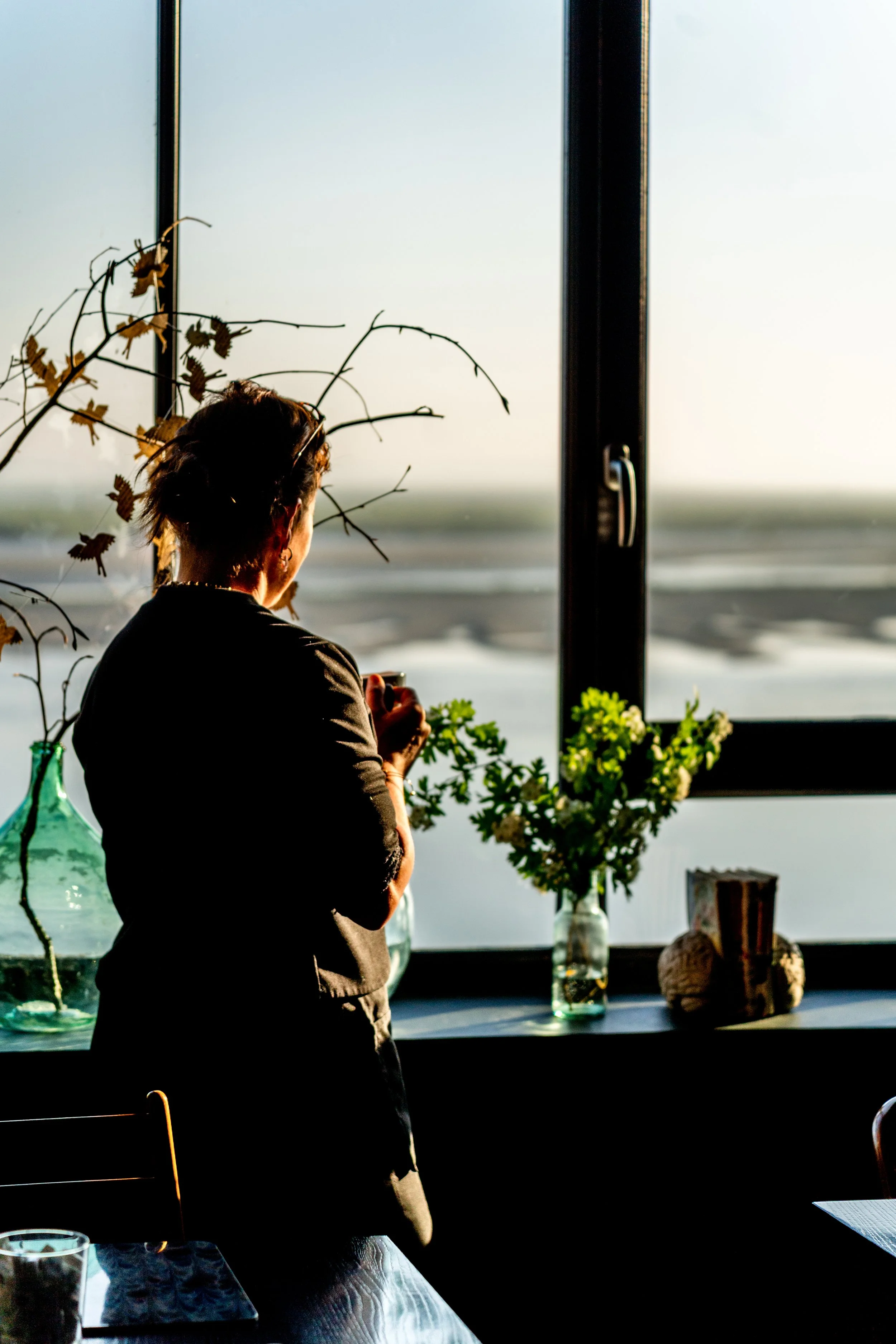 Une femme regardant par une fenêtre, entourée de vases avec des fleurs et des branches séchées, avec la lumière du soleil entrant dans la pièce.